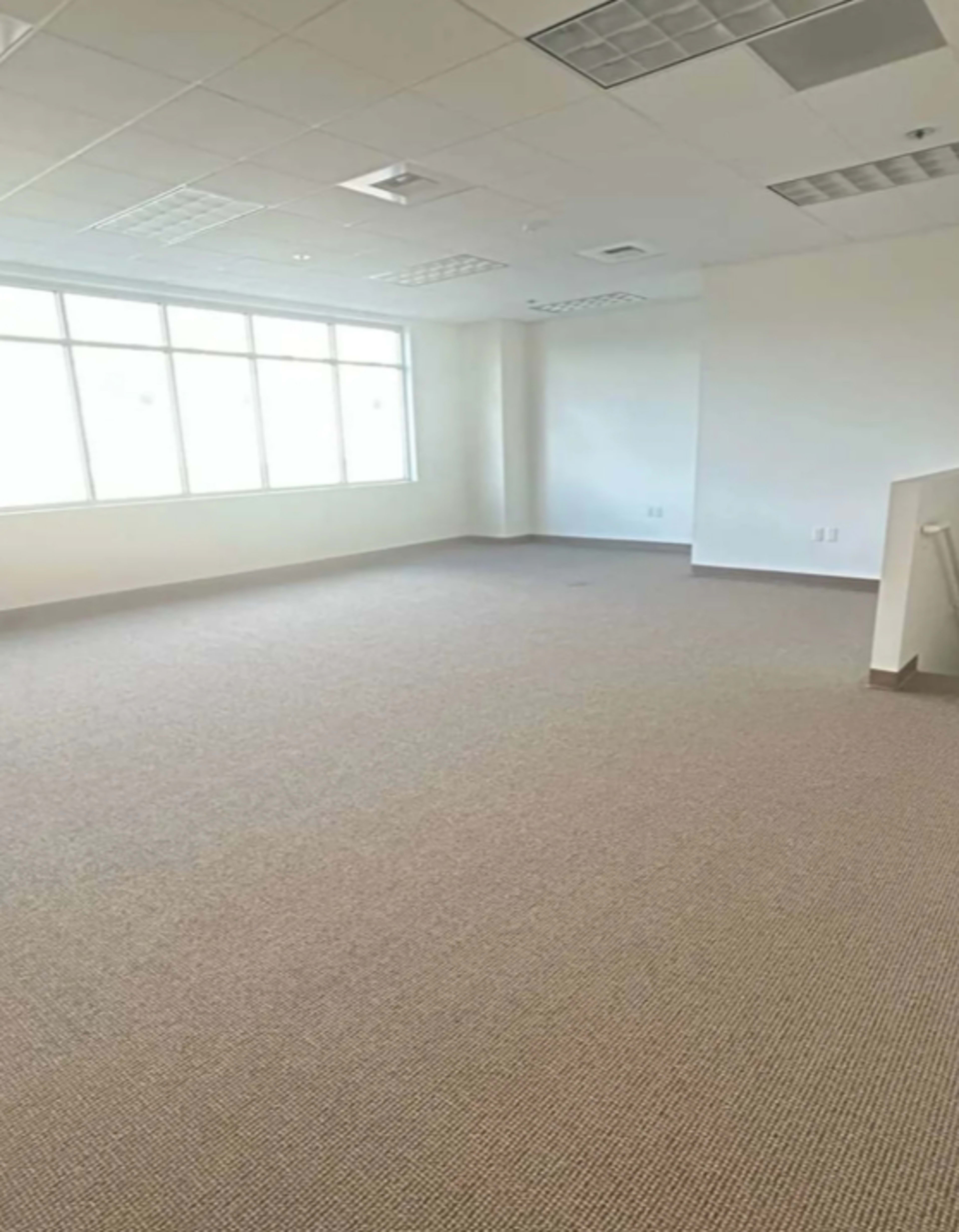 The image shows a spacious, empty office room with large windows allowing natural light to enter.