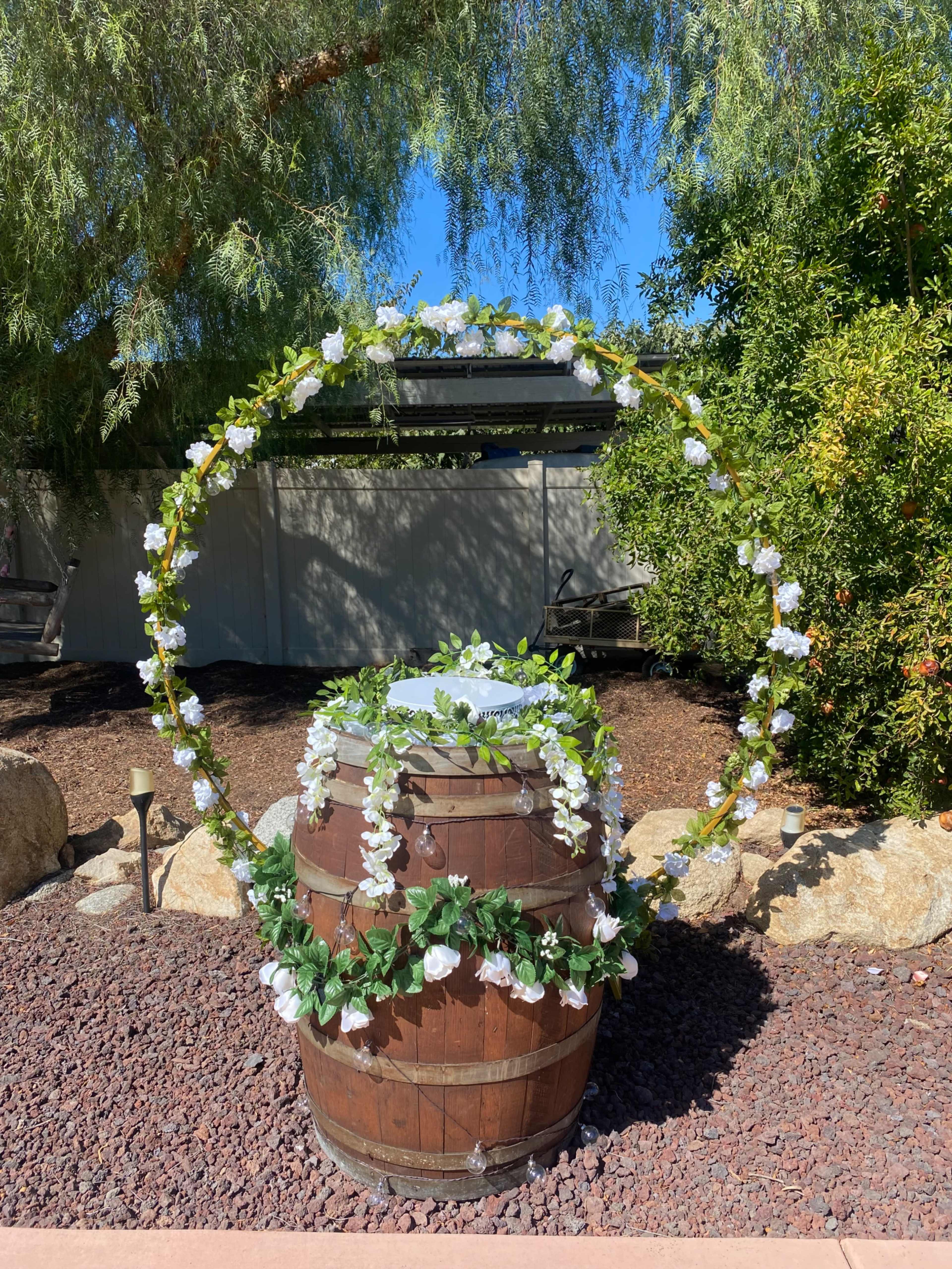 A wooden barrel is adorned with a circular arrangement of green leaves and white flowers, set against a backdrop of trees and a clear blue sky.