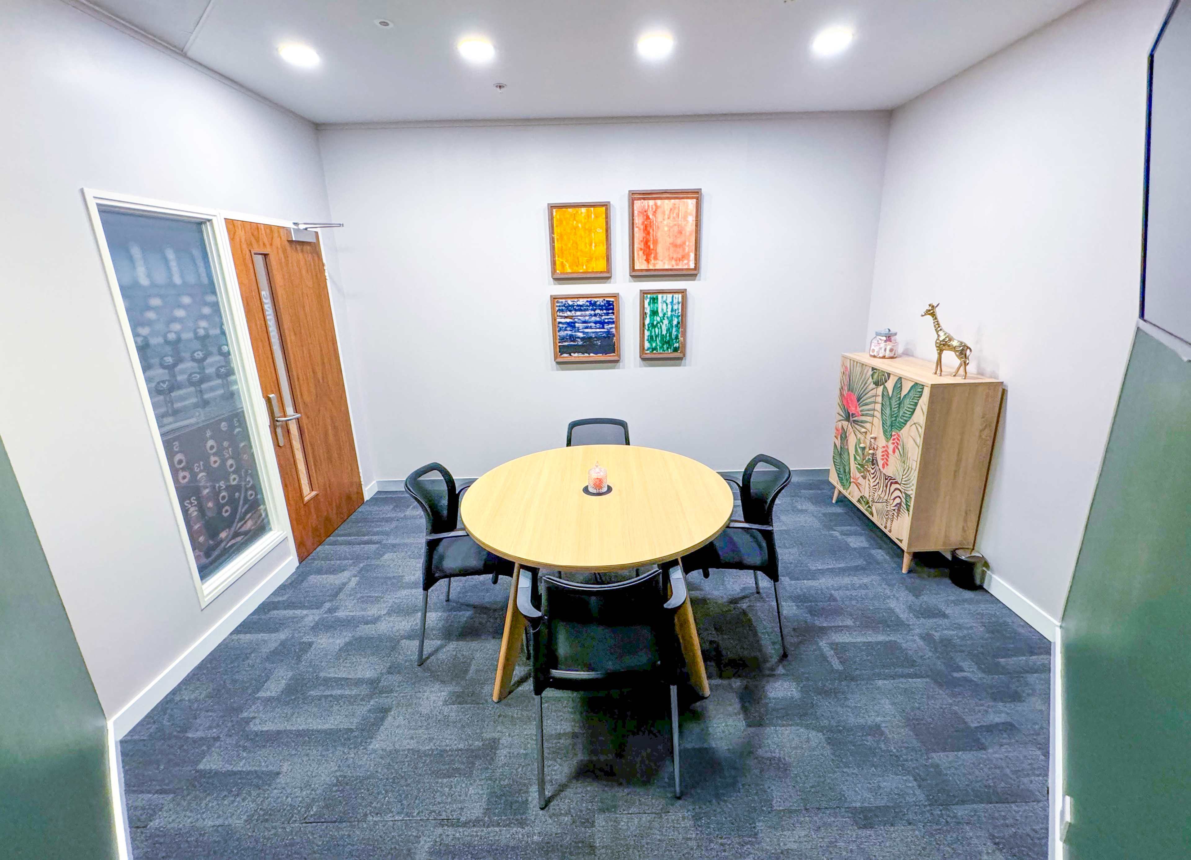 A small meeting room features a round wooden table surrounded by six chairs, with colorful artwork on the walls and a cabinet in the corner.