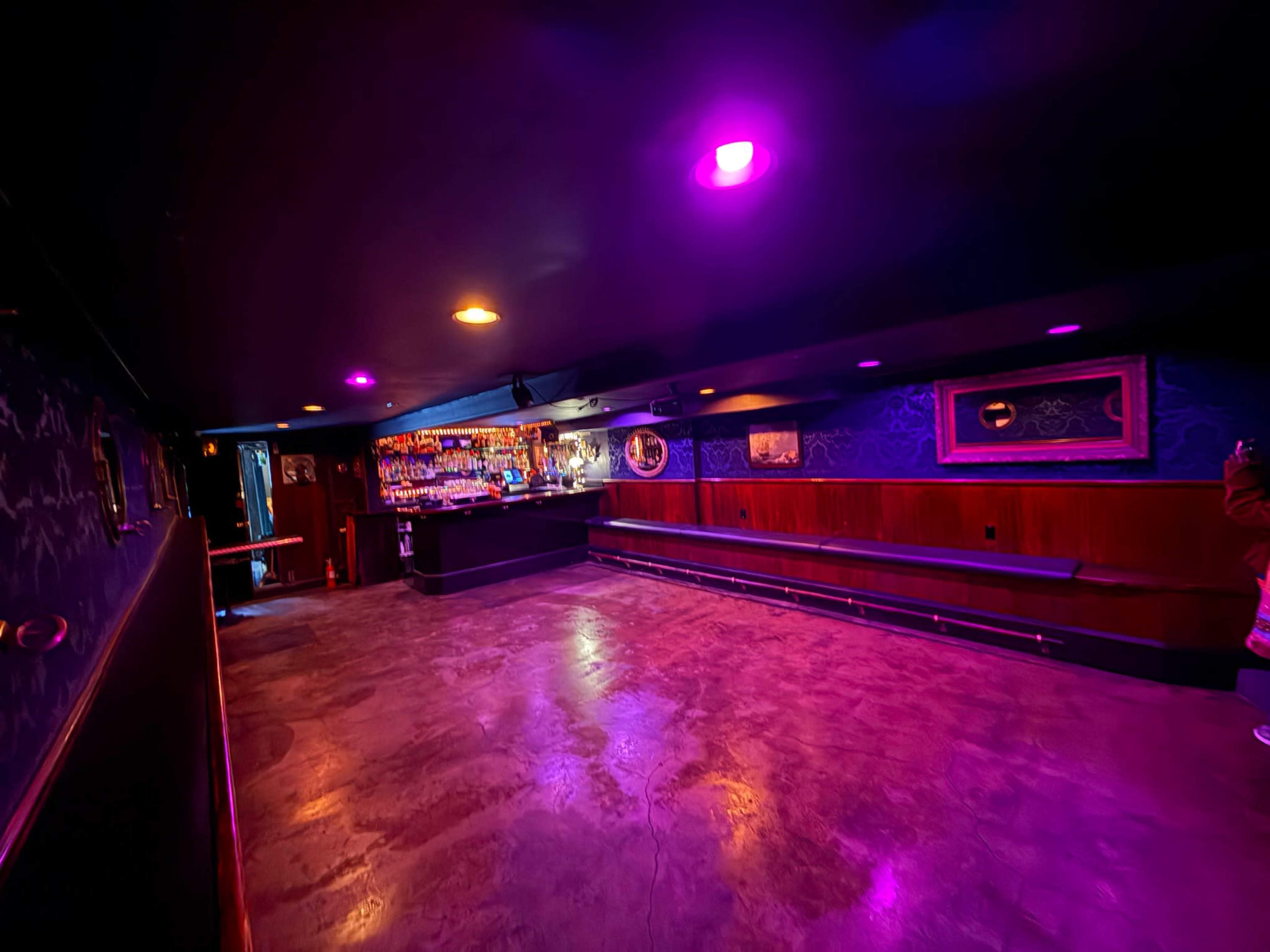 The image shows a dimly lit bar with purple and orange lights, featuring a long bench along the wall and a bar stocked with drinks in the background.