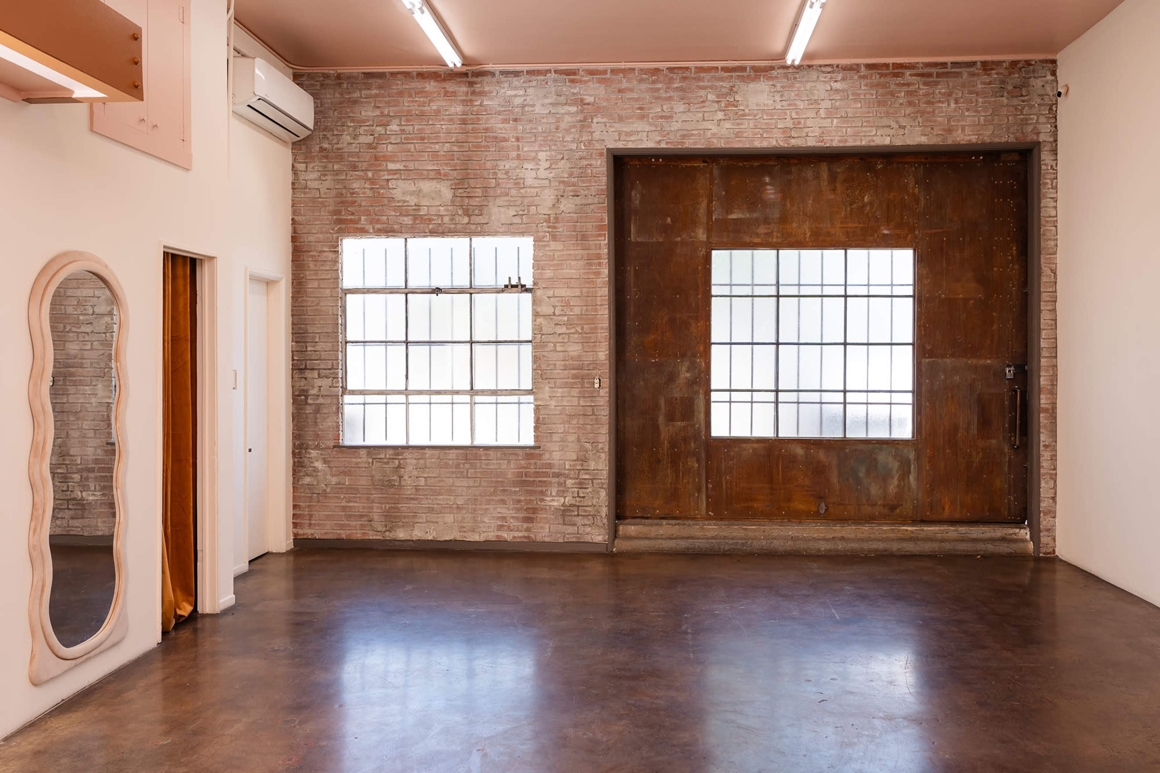 The image shows an interior space with exposed brick walls, a large rusted sliding door, and two windows with metal grids, along with a decorative mirror.