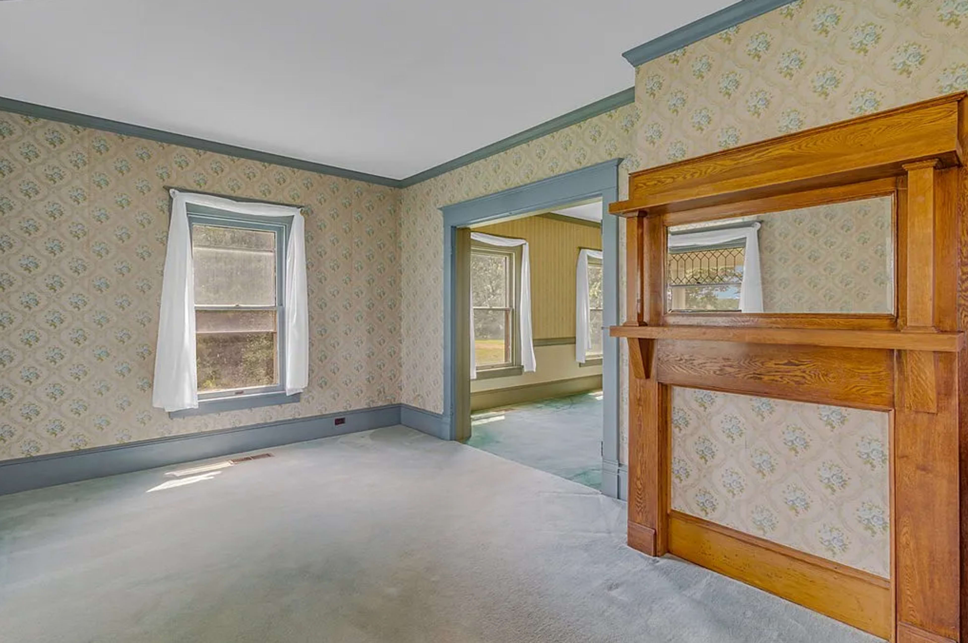 The image shows a living room with patterned wallpaper, two windows allowing natural light, and a wooden mantle piece.