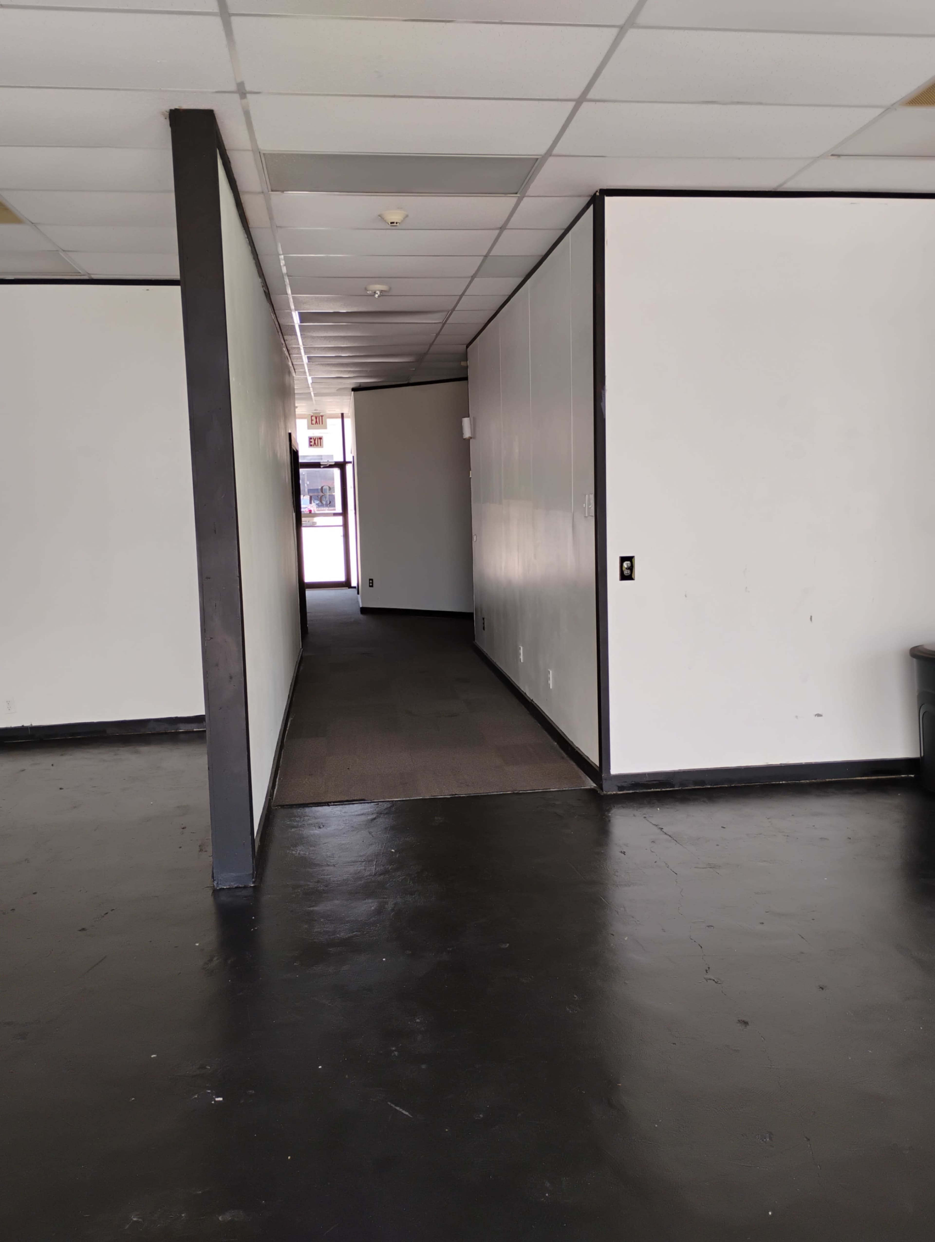 The image shows a vacant interior space with white walls, a dark floor, and a hallway leading to another room.