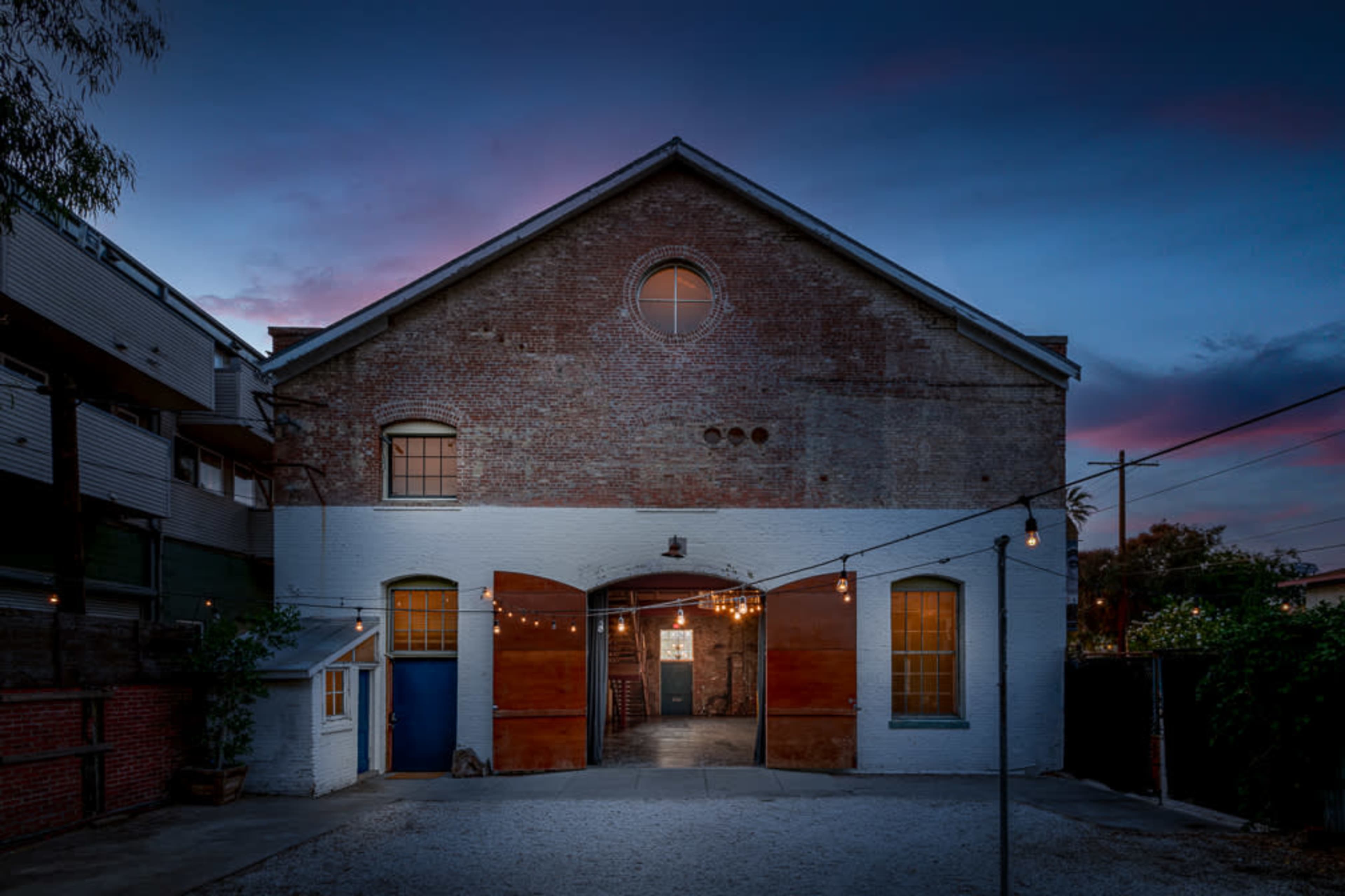 A large, brick building at dusk, featuring wooden double doors and string lights illuminating the entrance.