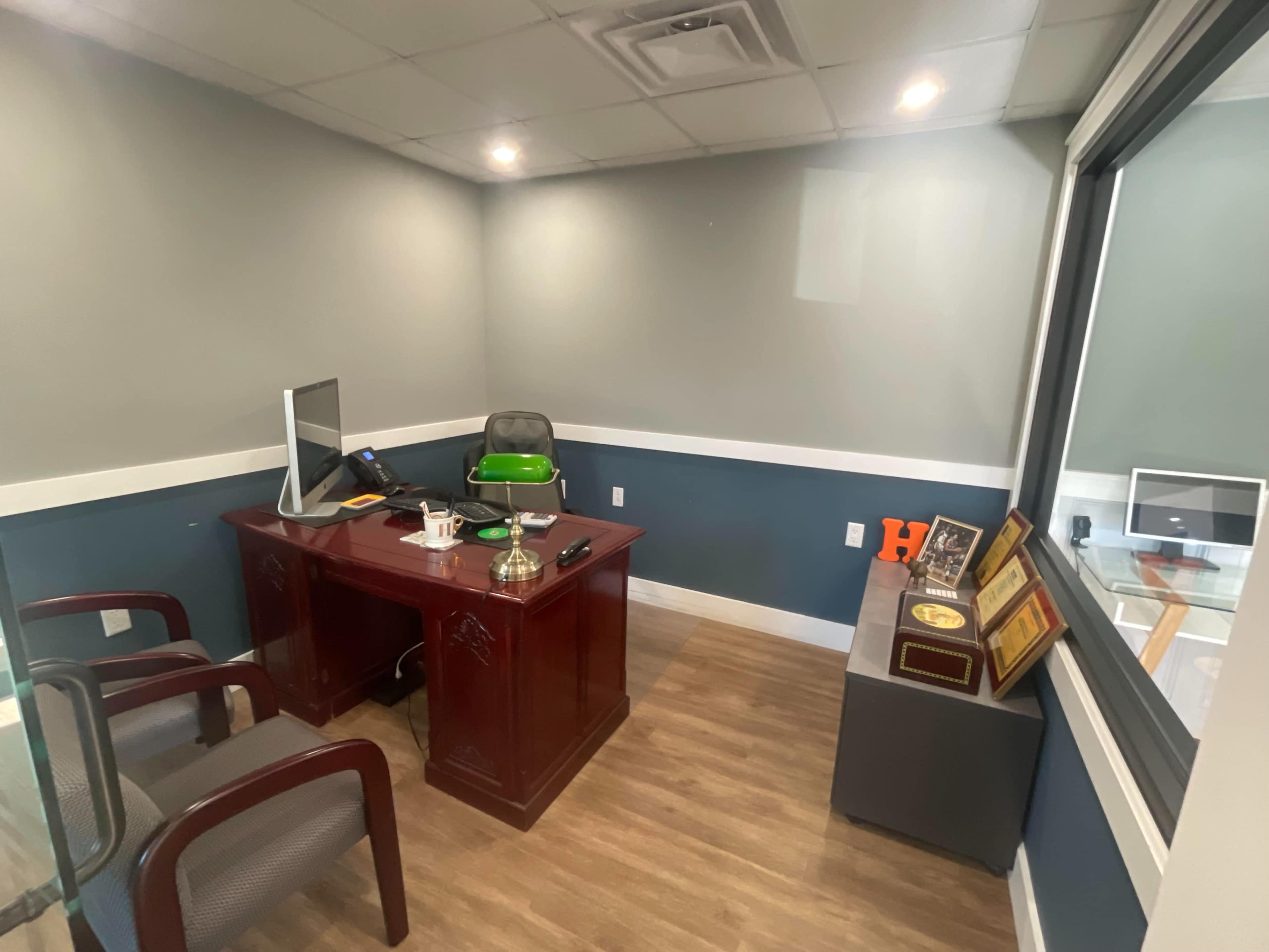 A small office features a wooden desk with a computer, a green chair, and two gray chairs beside it, along with a display of awards on a nearby cabinet.