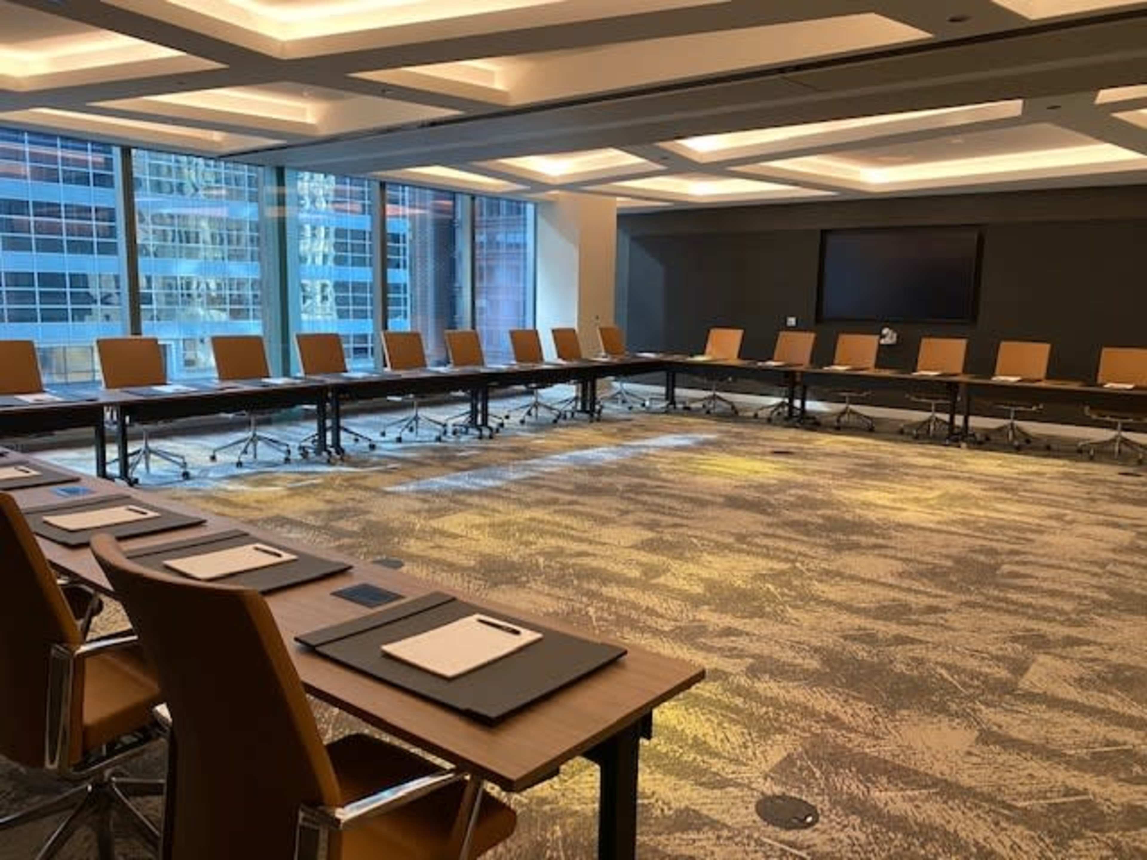The image shows a large conference room with a long rectangular table surrounded by chairs and large windows providing natural light.