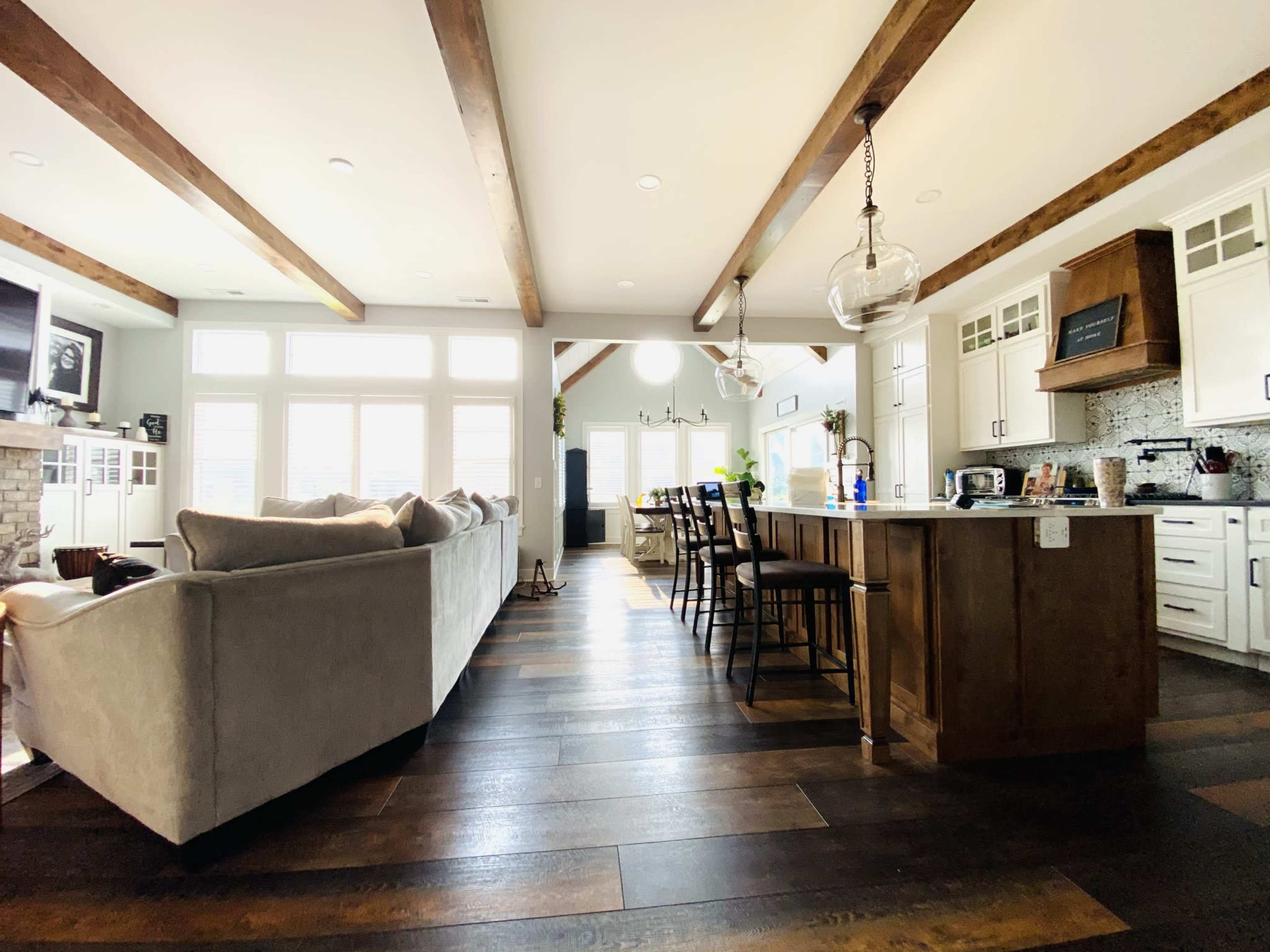 A modern kitchen and living area featuring wooden beams, a large sofa, a dining table with chairs, and ample natural light from several windows.