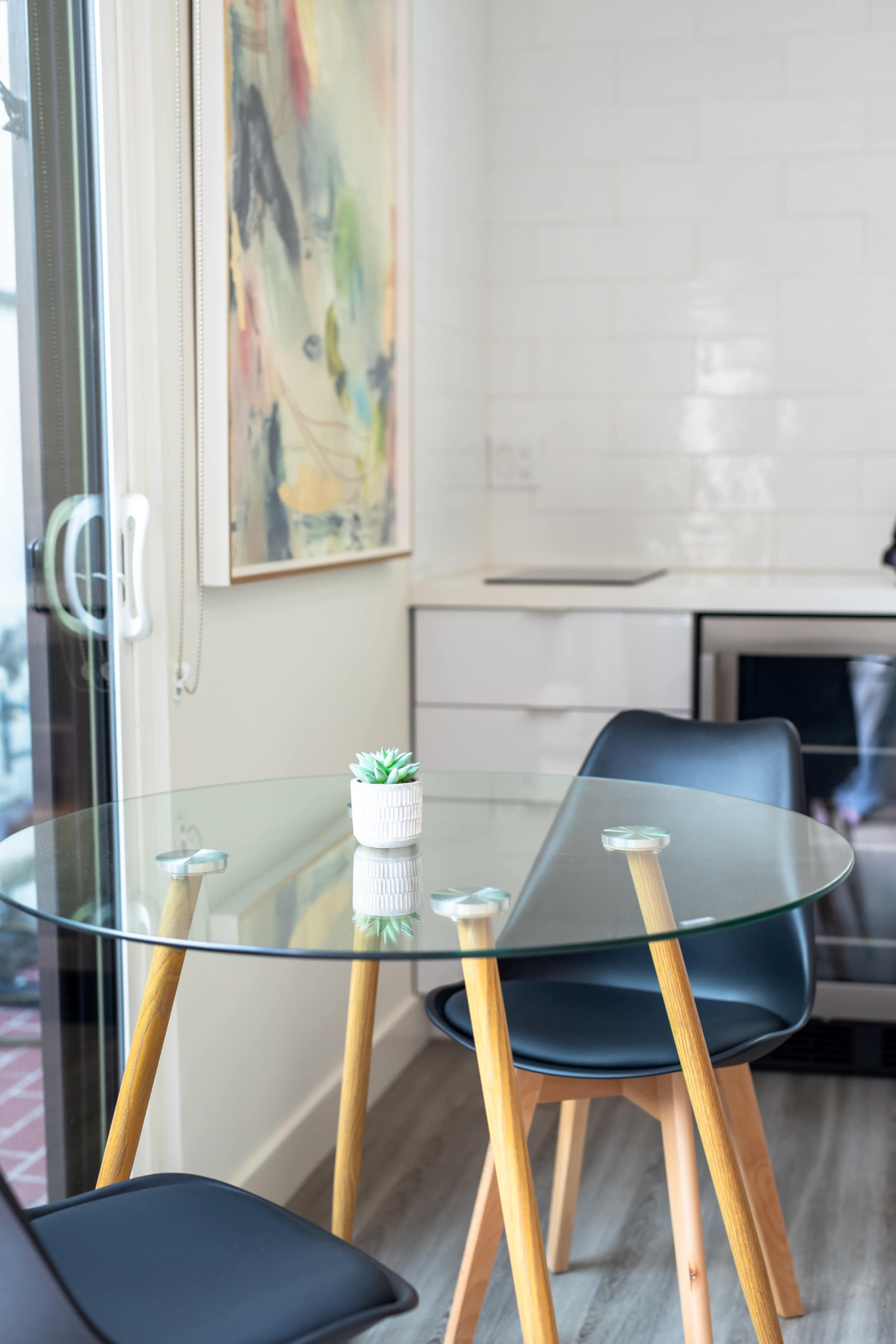 A round glass dining table with wooden legs is set in a bright, modern kitchen space, accompanied by two black chairs.