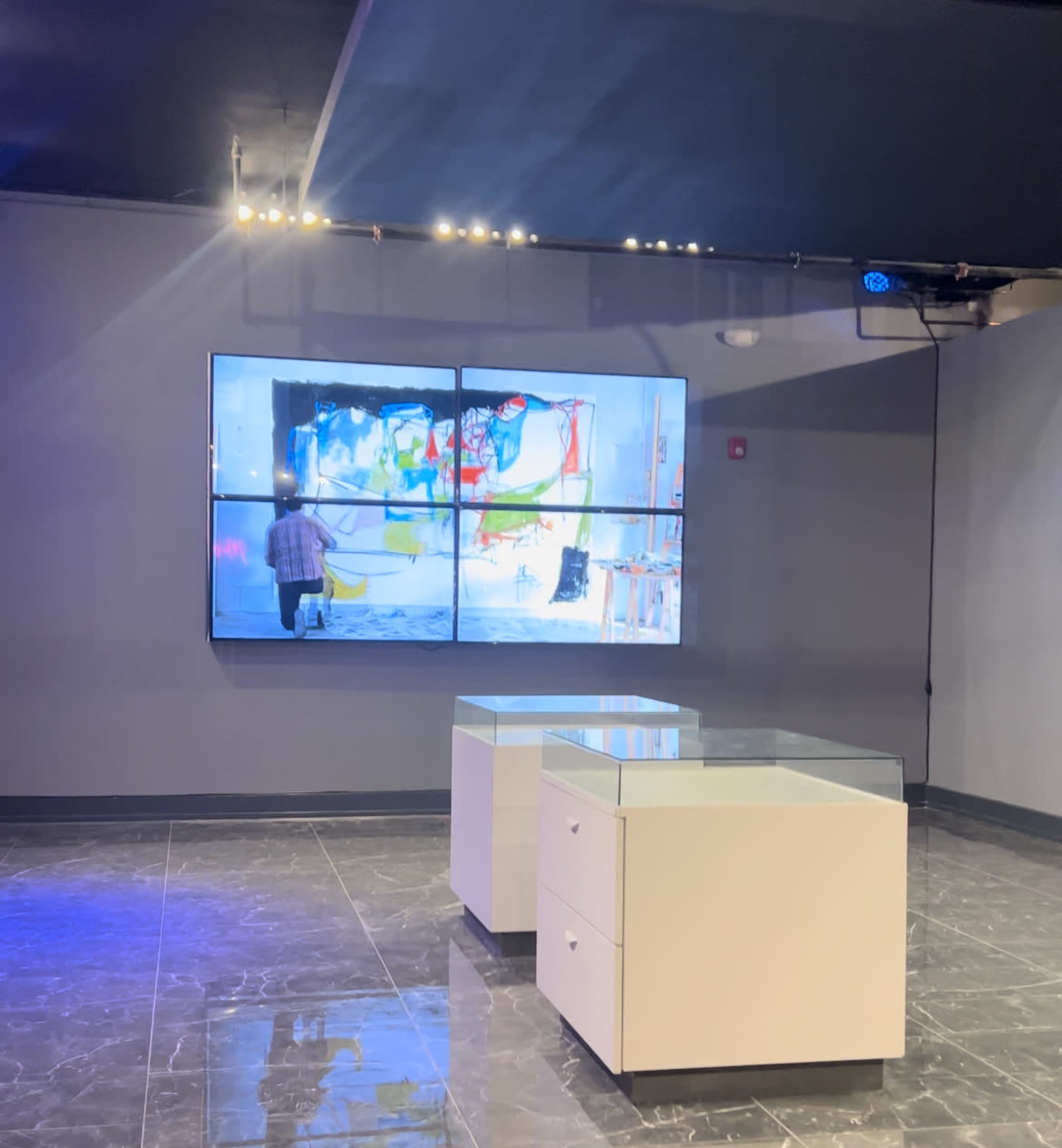 The image shows a modern gallery space featuring a large display screen showing an artist at work, with two white display tables in the foreground.