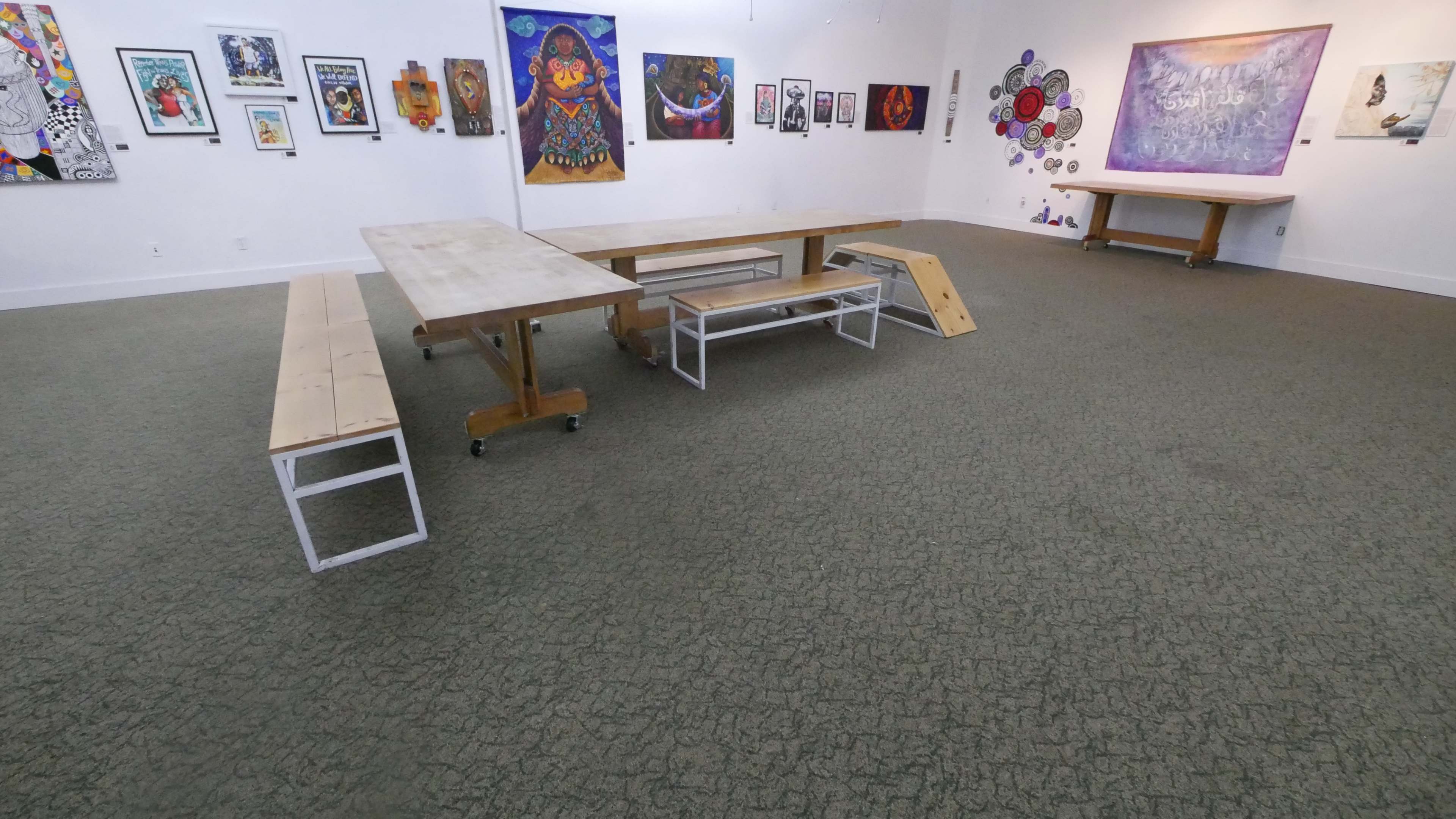 A spacious art gallery with wooden tables and benches arranged in the center, surrounded by colorful artwork on the walls.