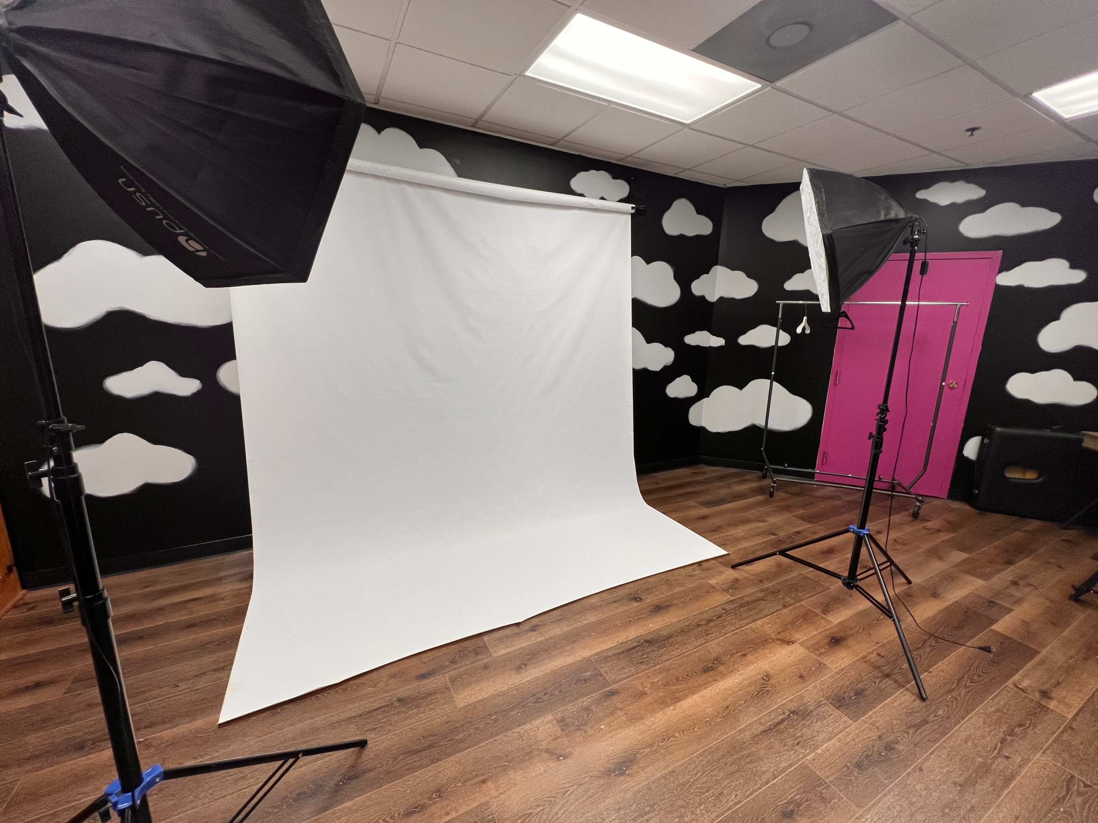 The image shows a photography studio with a white backdrop, two softbox lights, and walls decorated with cloud patterns.