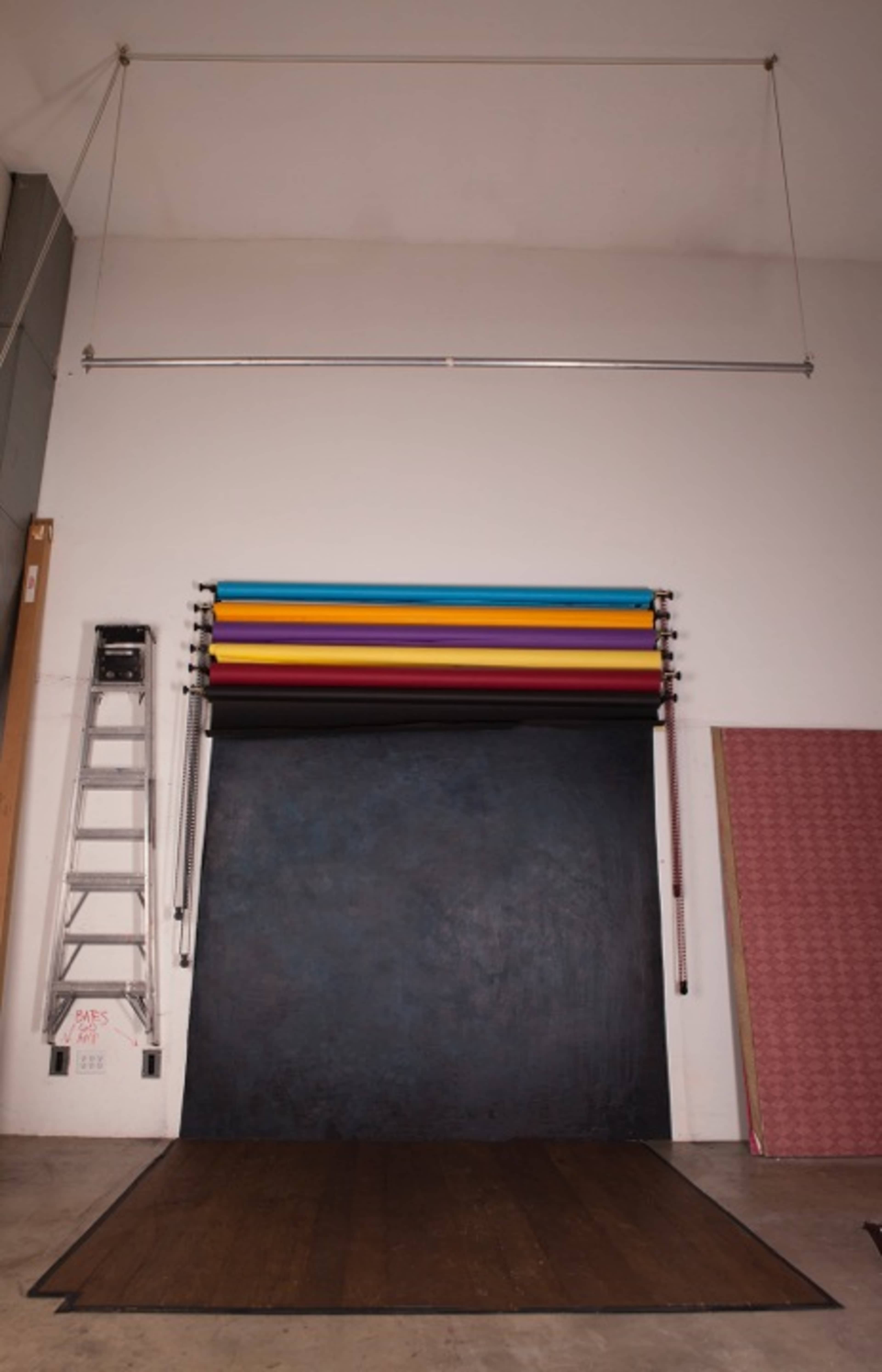 The image shows a photography studio setup with a large black backdrop, a ladder, and several colored backdrops hanging from a rod on the wall.