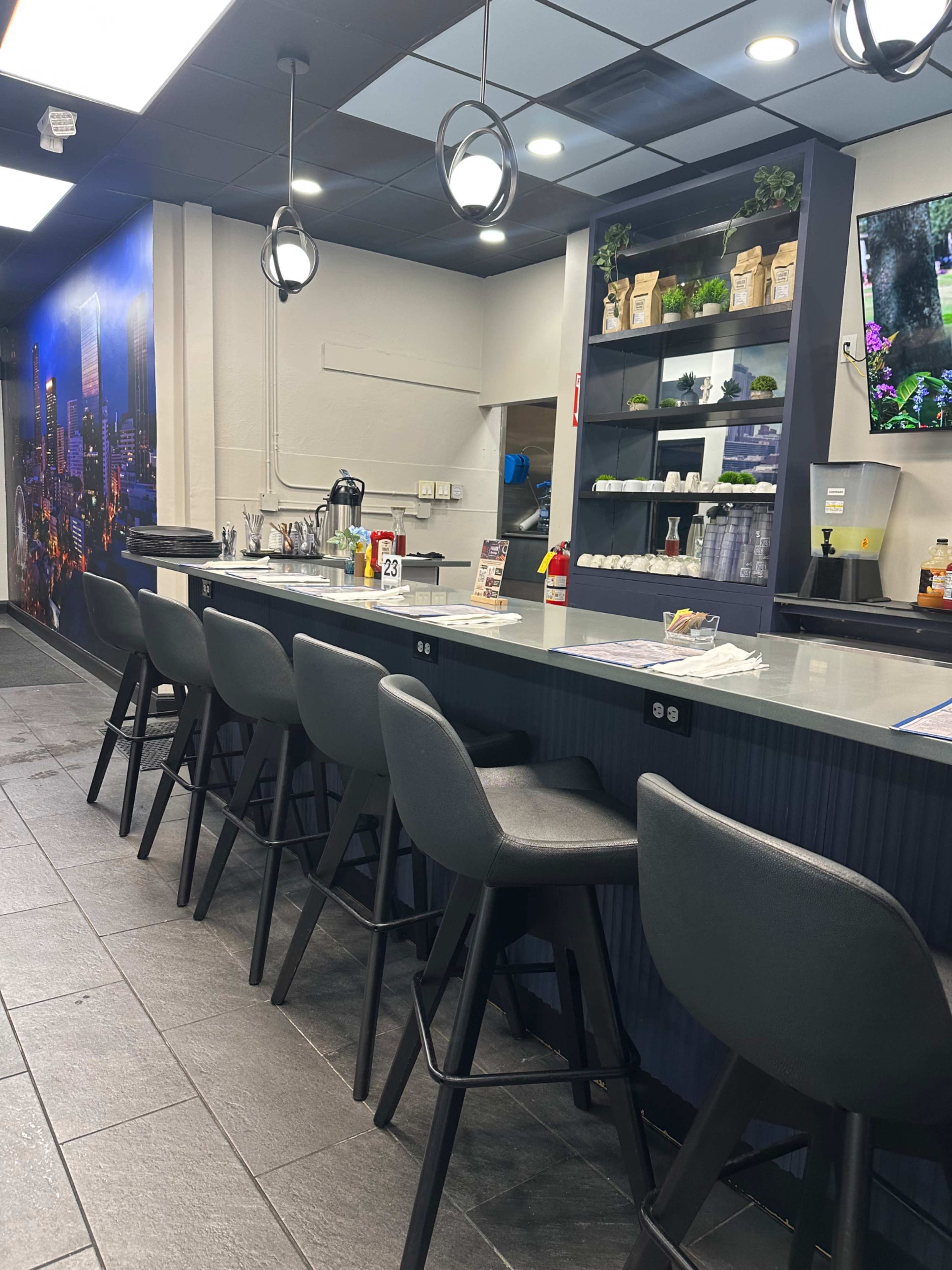 The image shows a modern counter area in a restaurant with high stools, a wall displaying cityscape images, and food items neatly arranged on shelves.