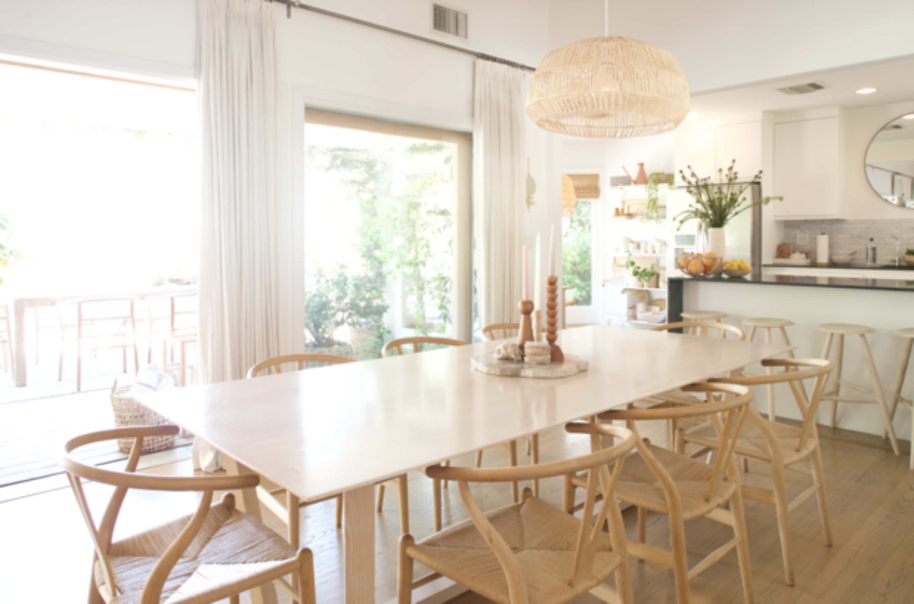 A spacious dining area features a large wooden table surrounded by woven chairs, illuminated by a hanging light fixture, with a view of an outdoor space beyond glass doors.