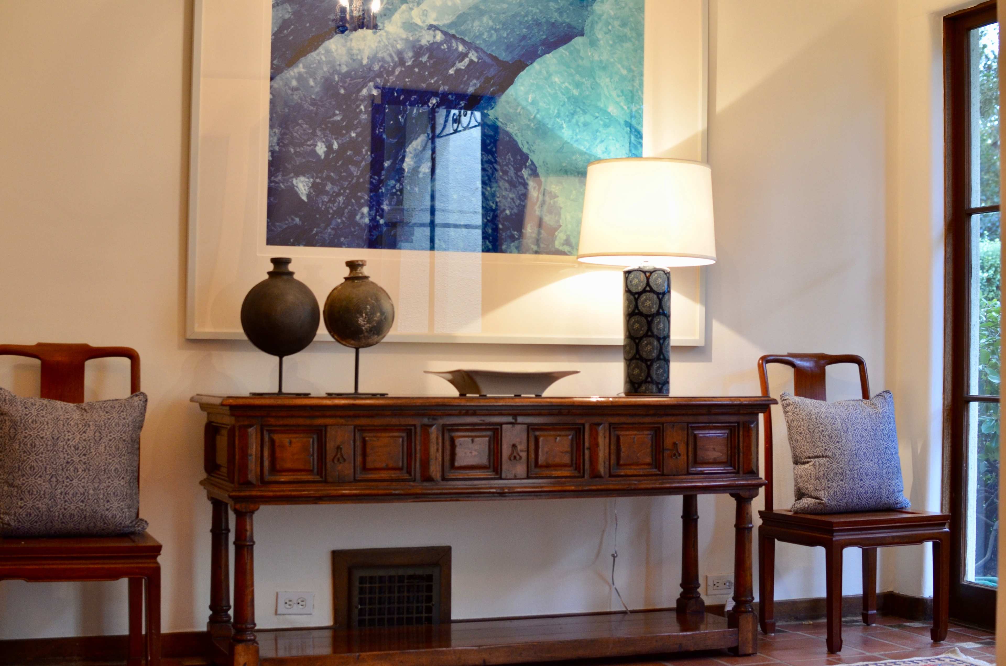 A wooden console table with decorative items and a lamp stands against a wall featuring a large abstract painting, flanked by two chairs with cushions.