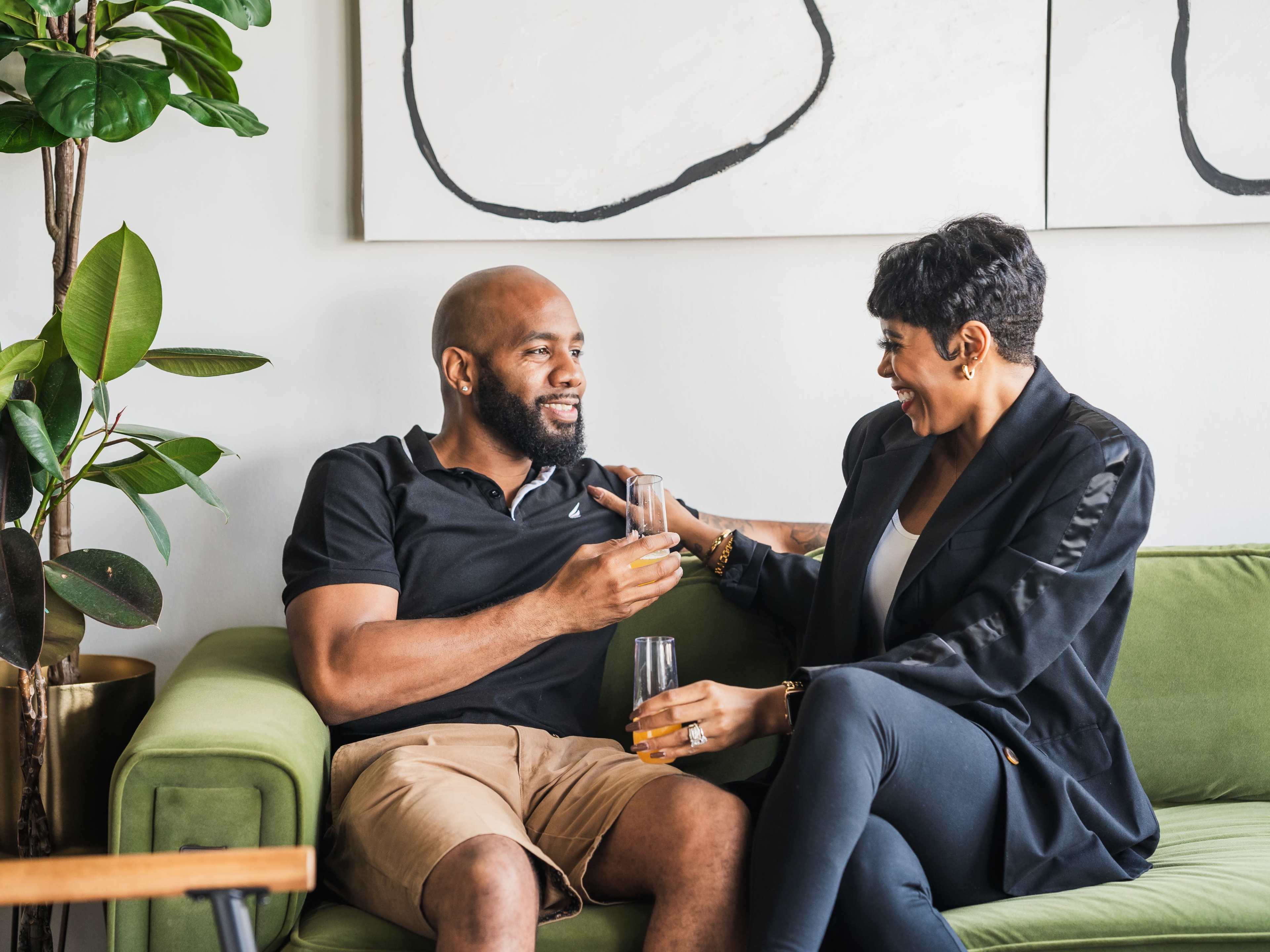 A man and woman sit together on a green sofa, laughing and enjoying drinks in a stylish living room.