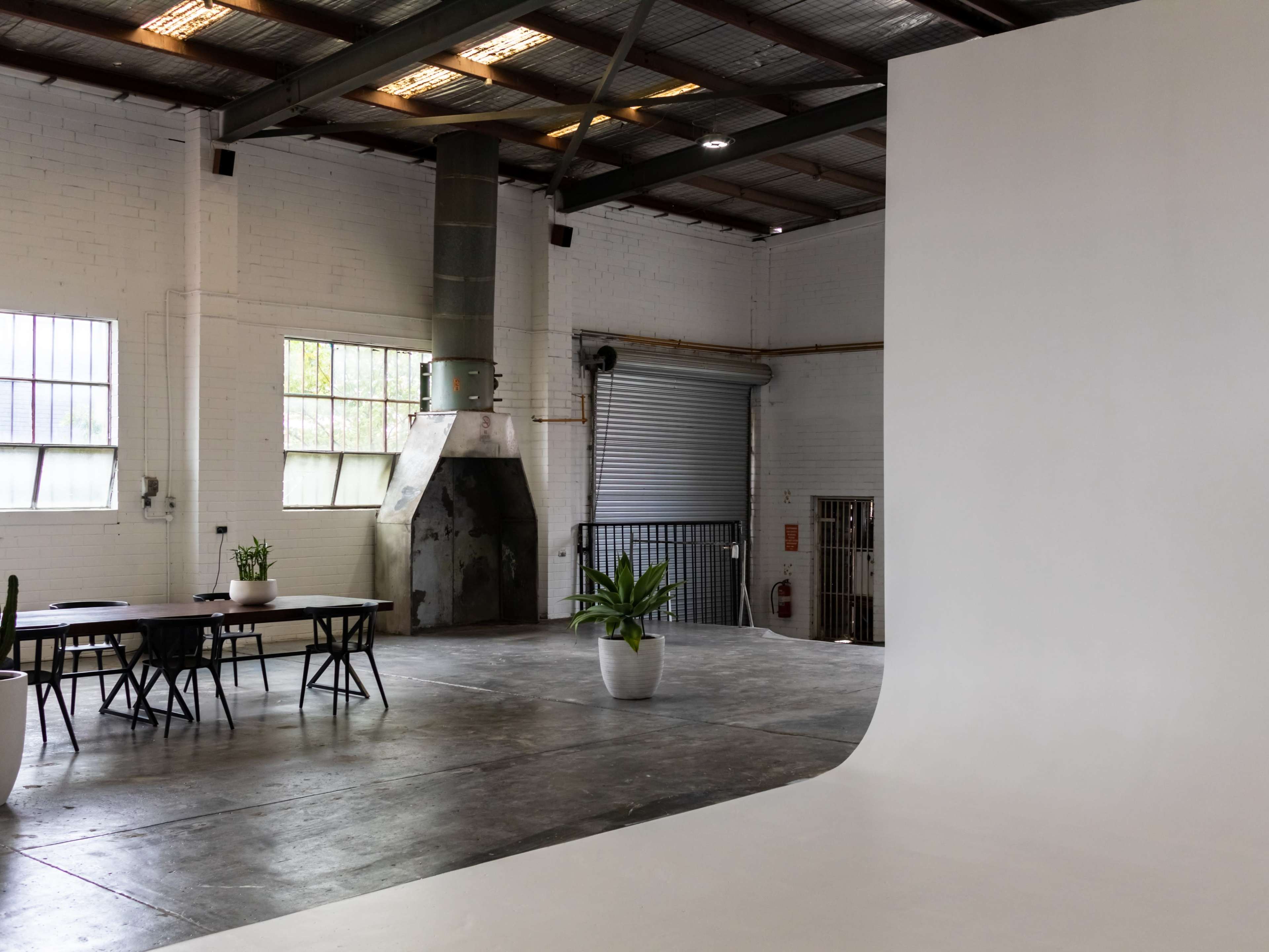 An industrial-style studio space with a large white backdrop, a table with chairs, and a prominent ventilation system.