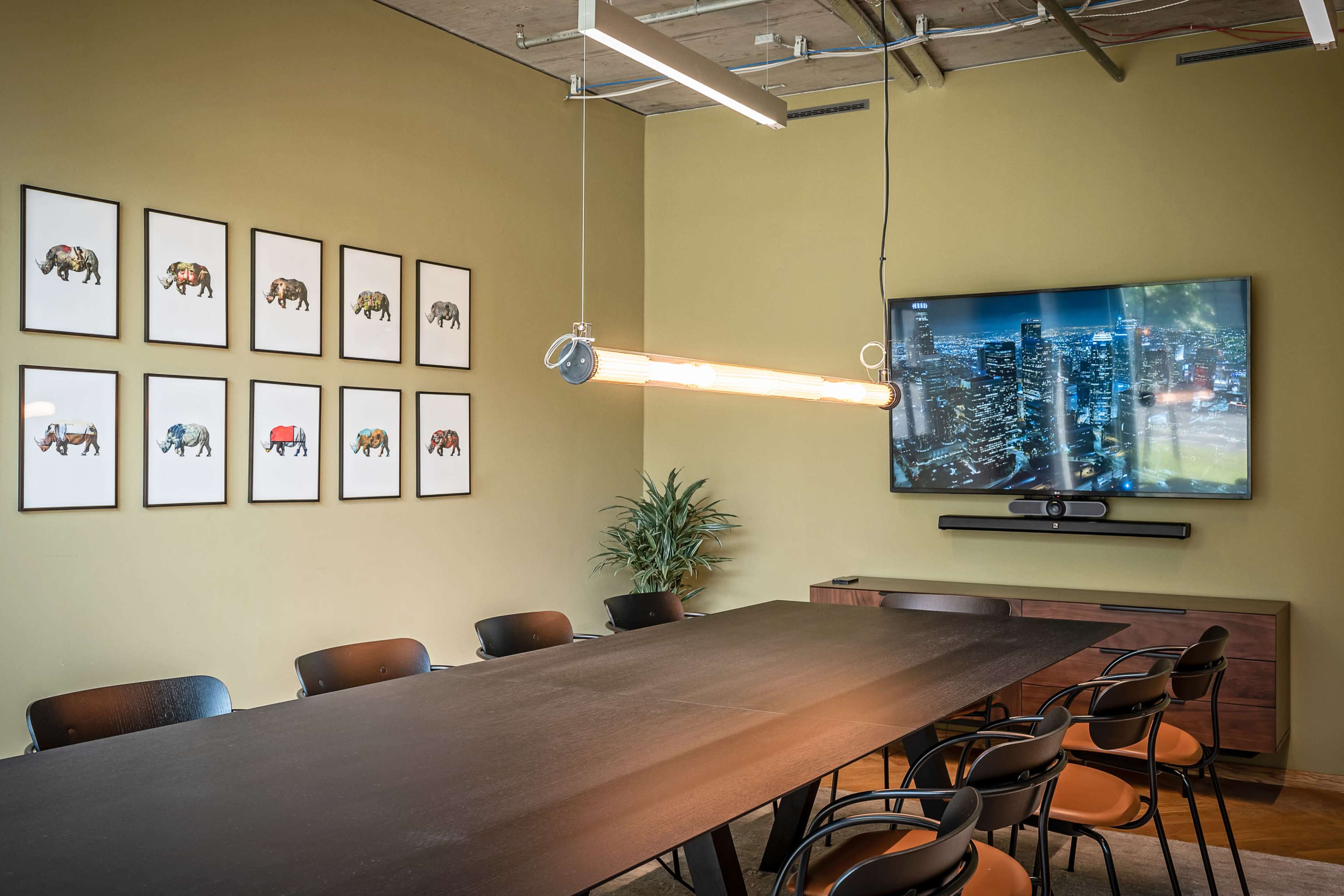 The image shows a meeting room featuring a long black table, black chairs, a wall with framed artwork of elephants, and a large television mounted on the opposite wall.