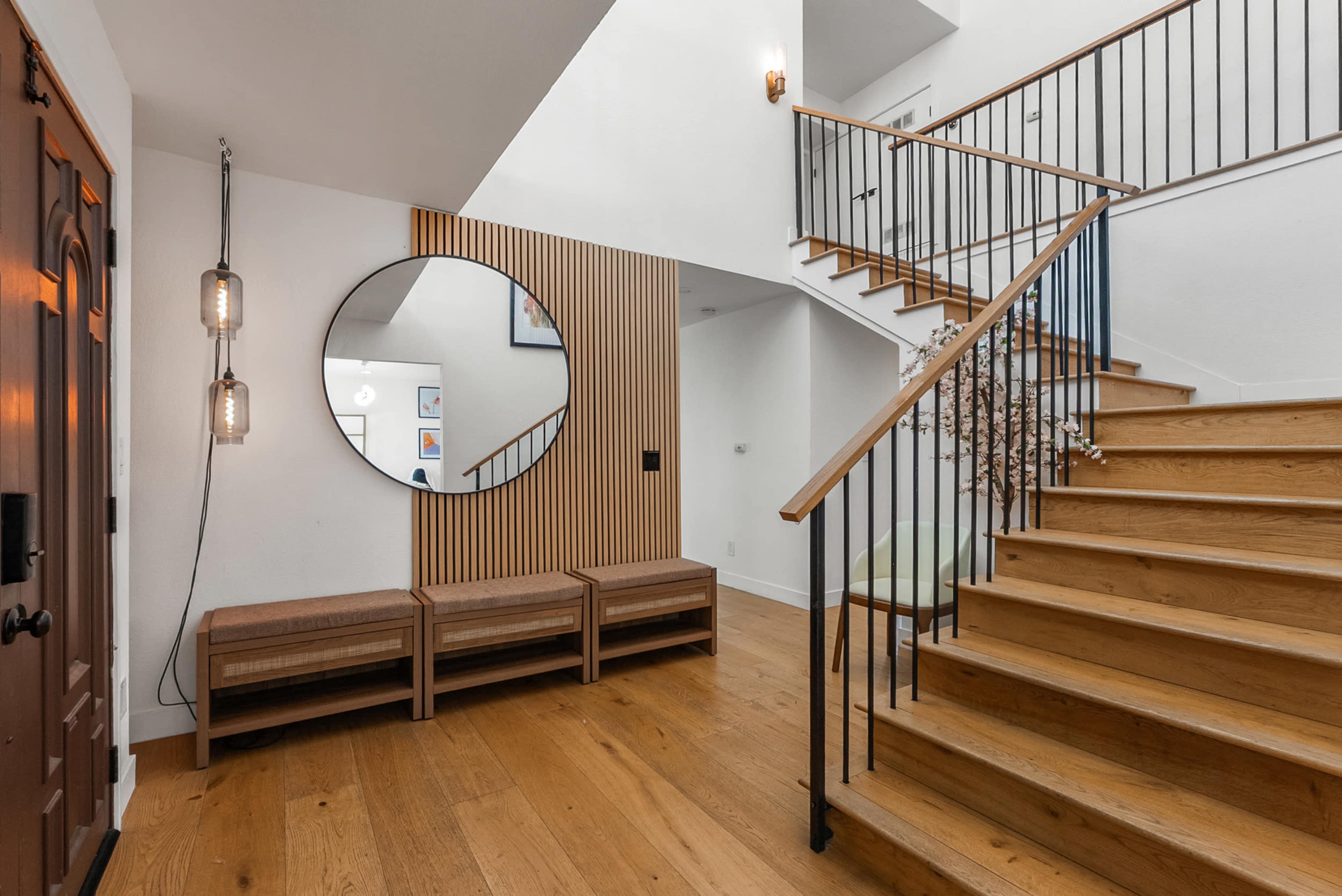 The image shows a modern entrance hall with wooden flooring, a staircase, a round mirror, and a bench area near the front door.