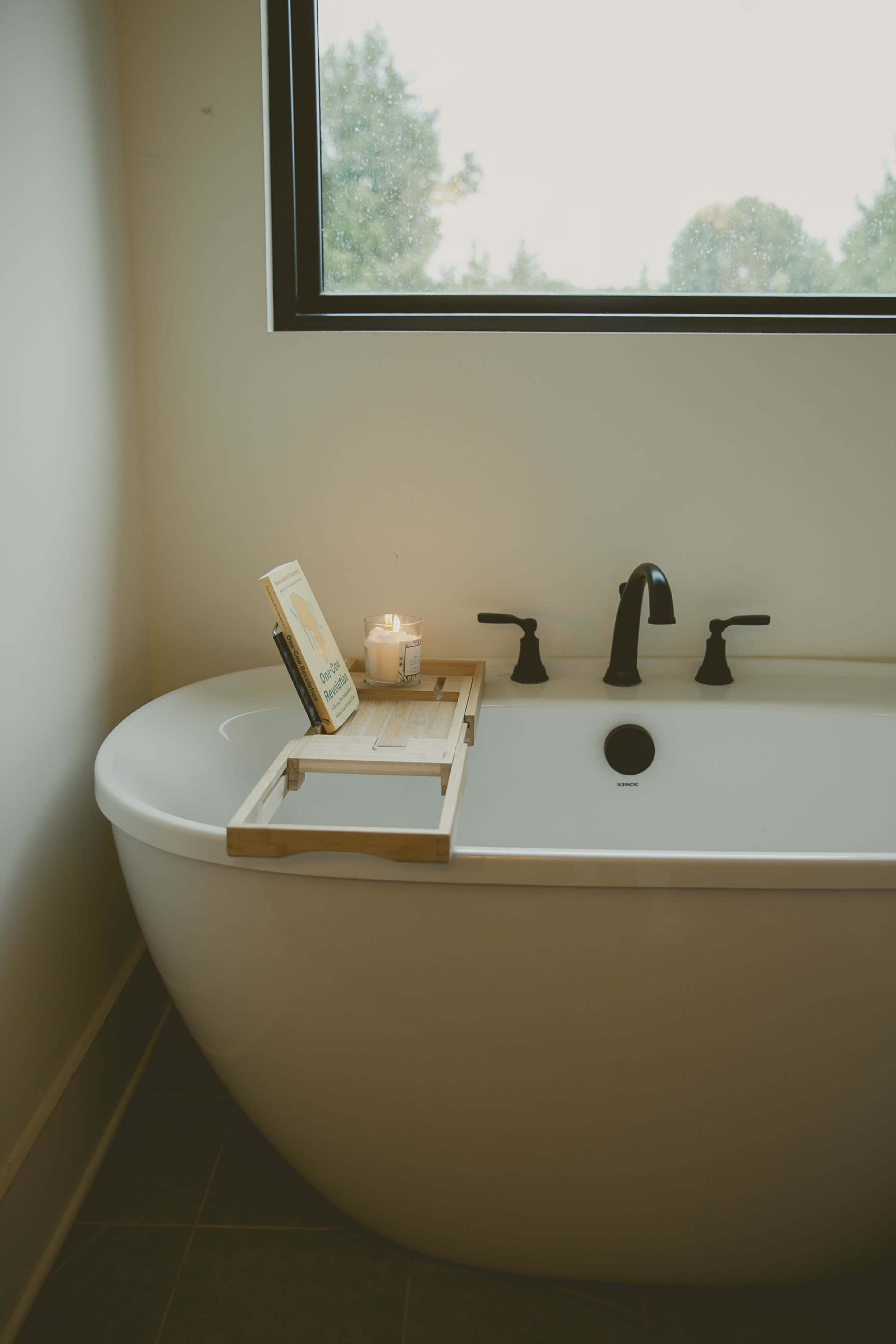 A modern bathroom features a freestanding oval bathtub with a wooden tray holding a book and a candle beside a window that offers a view of greenery.