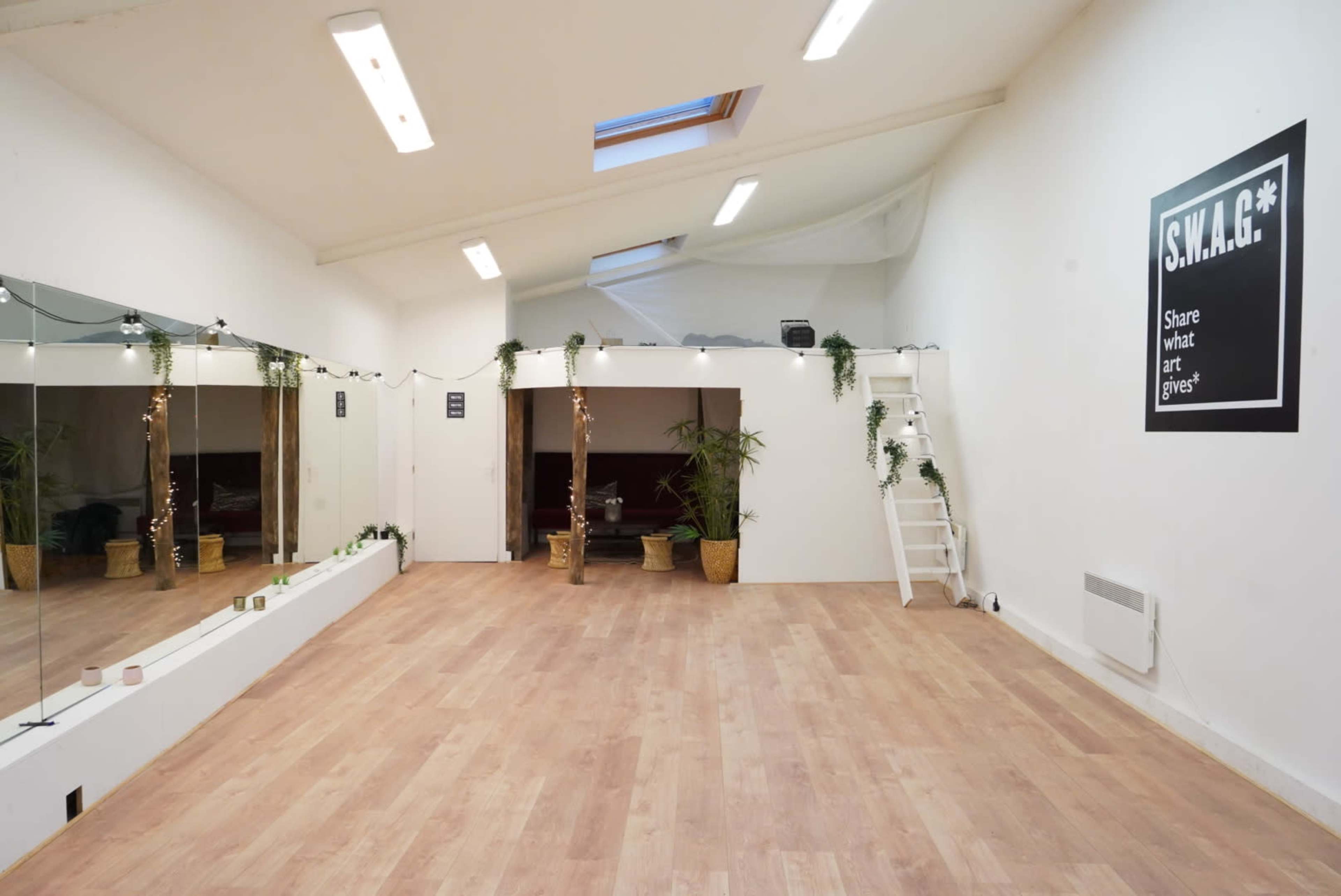 The interior of a spacious, well-lit studio features a mirrored wall, potted plants, and a sign that reads "S.W.A.G.*" on one side.
