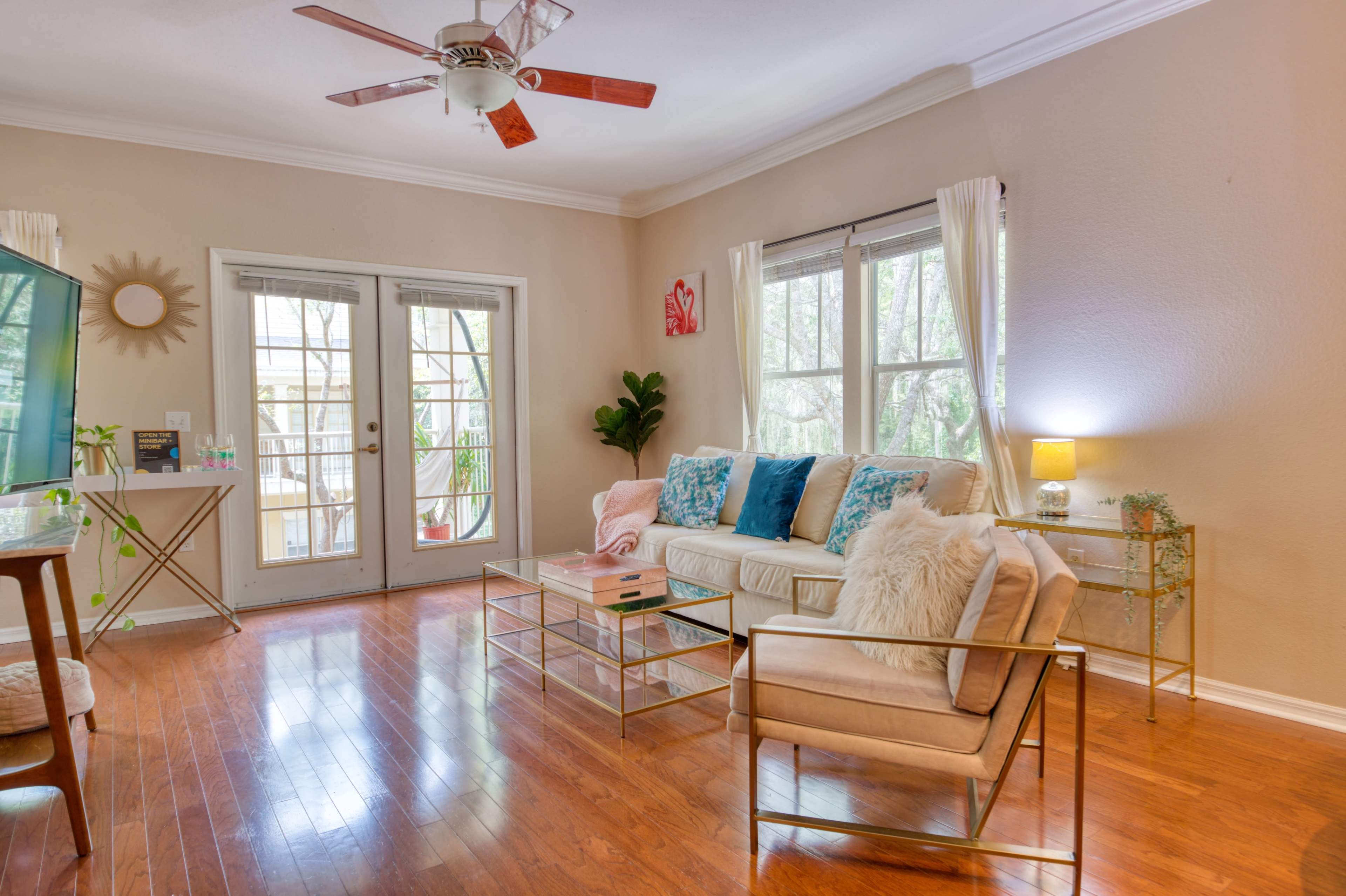 The room features a light-colored sofa with decorative pillows, a glass coffee table, and large windows that open to a balcony, illuminated by natural light.