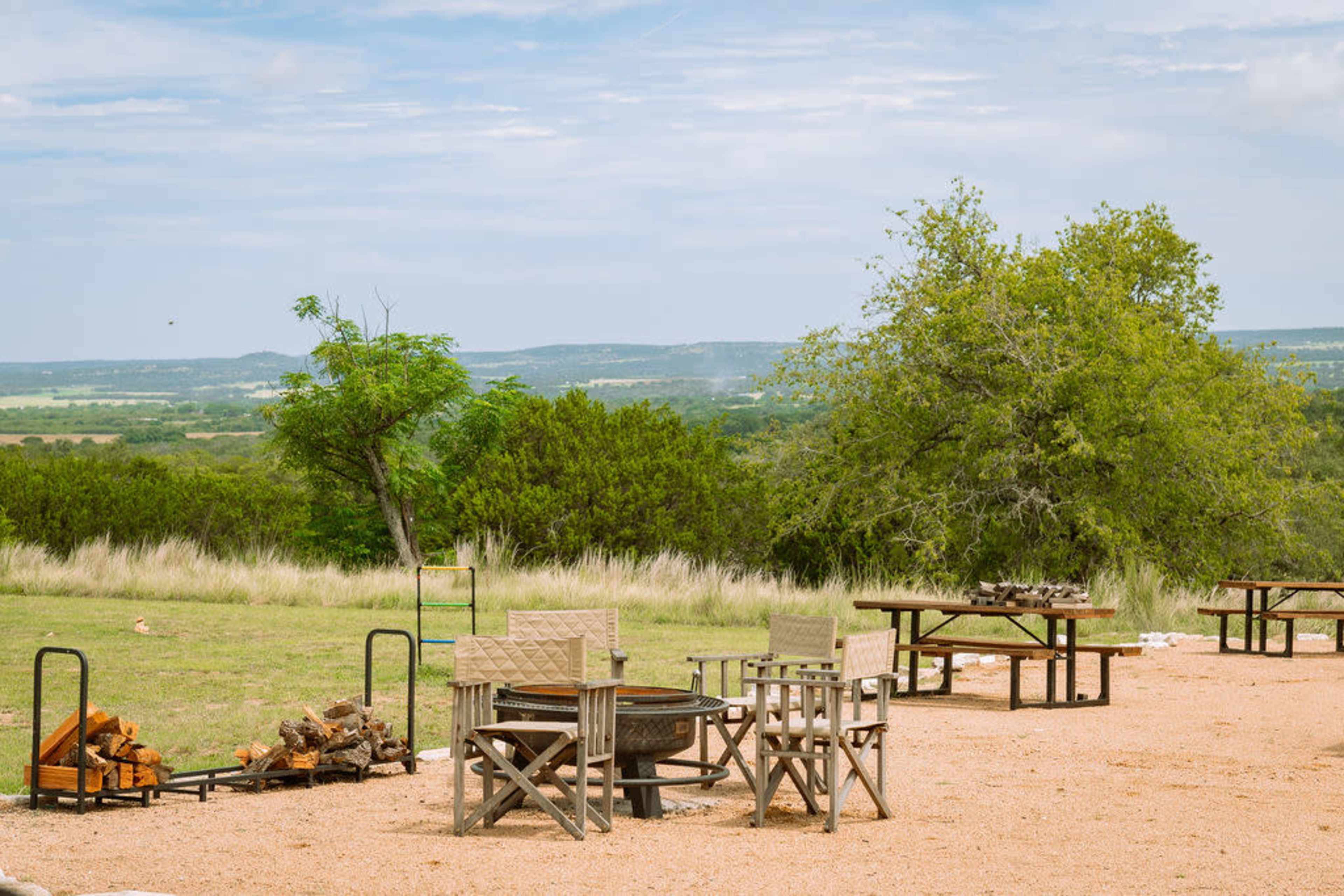 The scene features a spacious outdoor area with wooden tables and chairs, surrounded by grassy fields and distant hills under a cloudy sky.