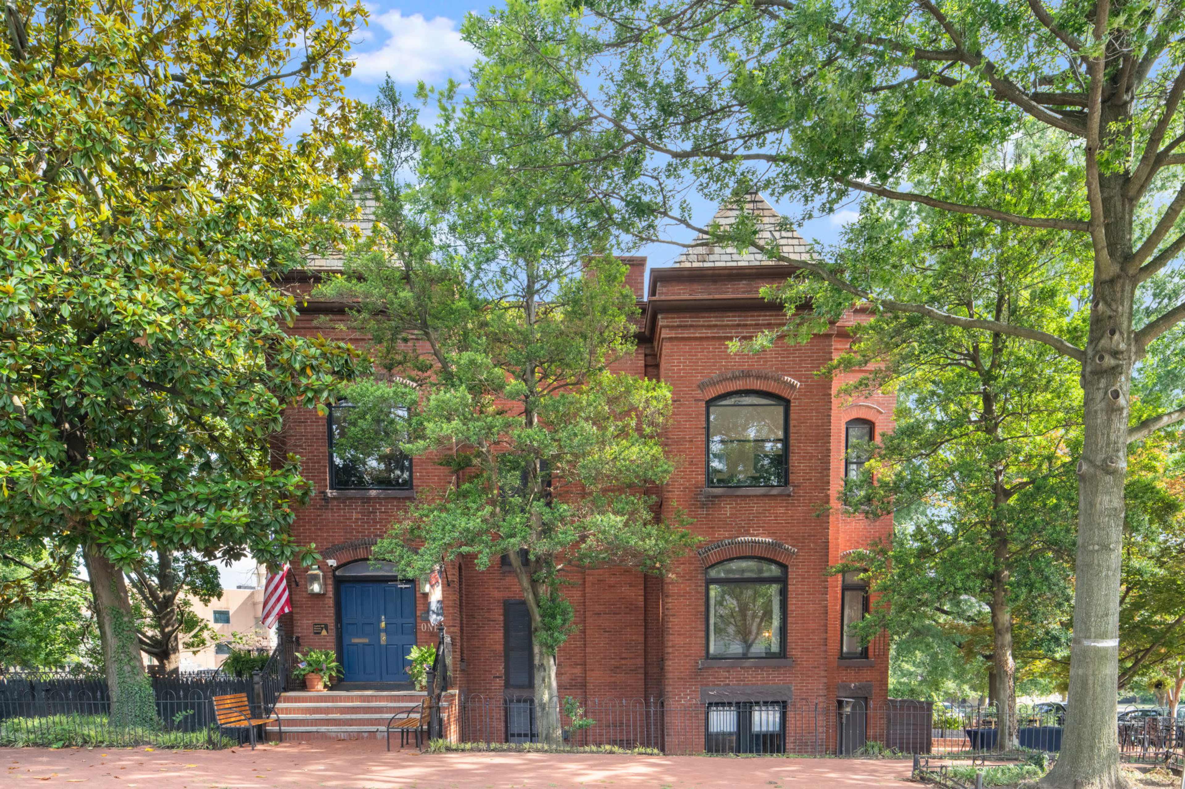 The image shows a two-story brick house with large windows, surrounded by trees and a small garden.
