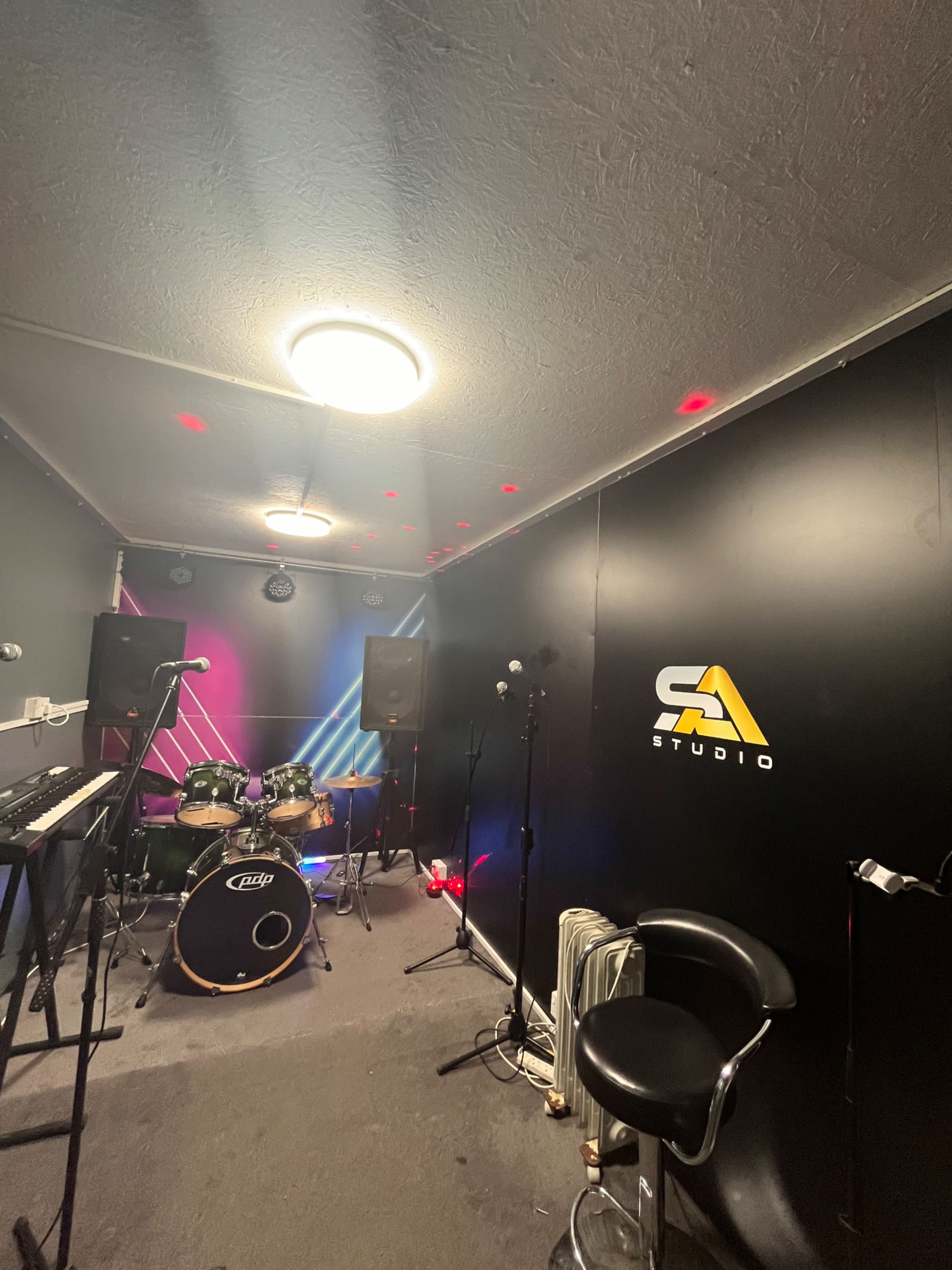 The image shows a small music studio equipped with a drum set, keyboards, and microphones, featuring colorful lights and a logo on the wall.