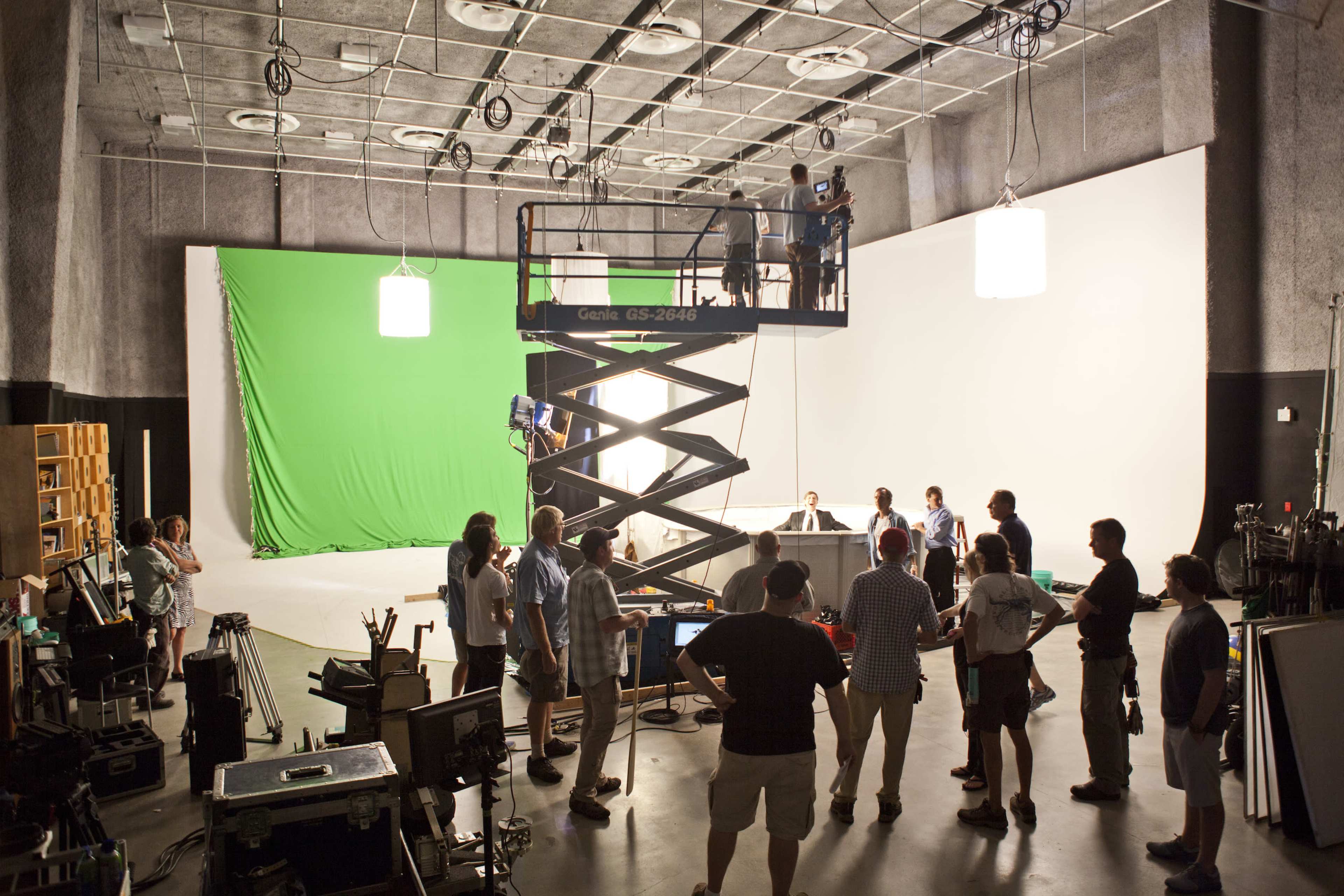 A film crew is set up in a studio, with a scissor lift in the center and a green screen backdrop on one side.