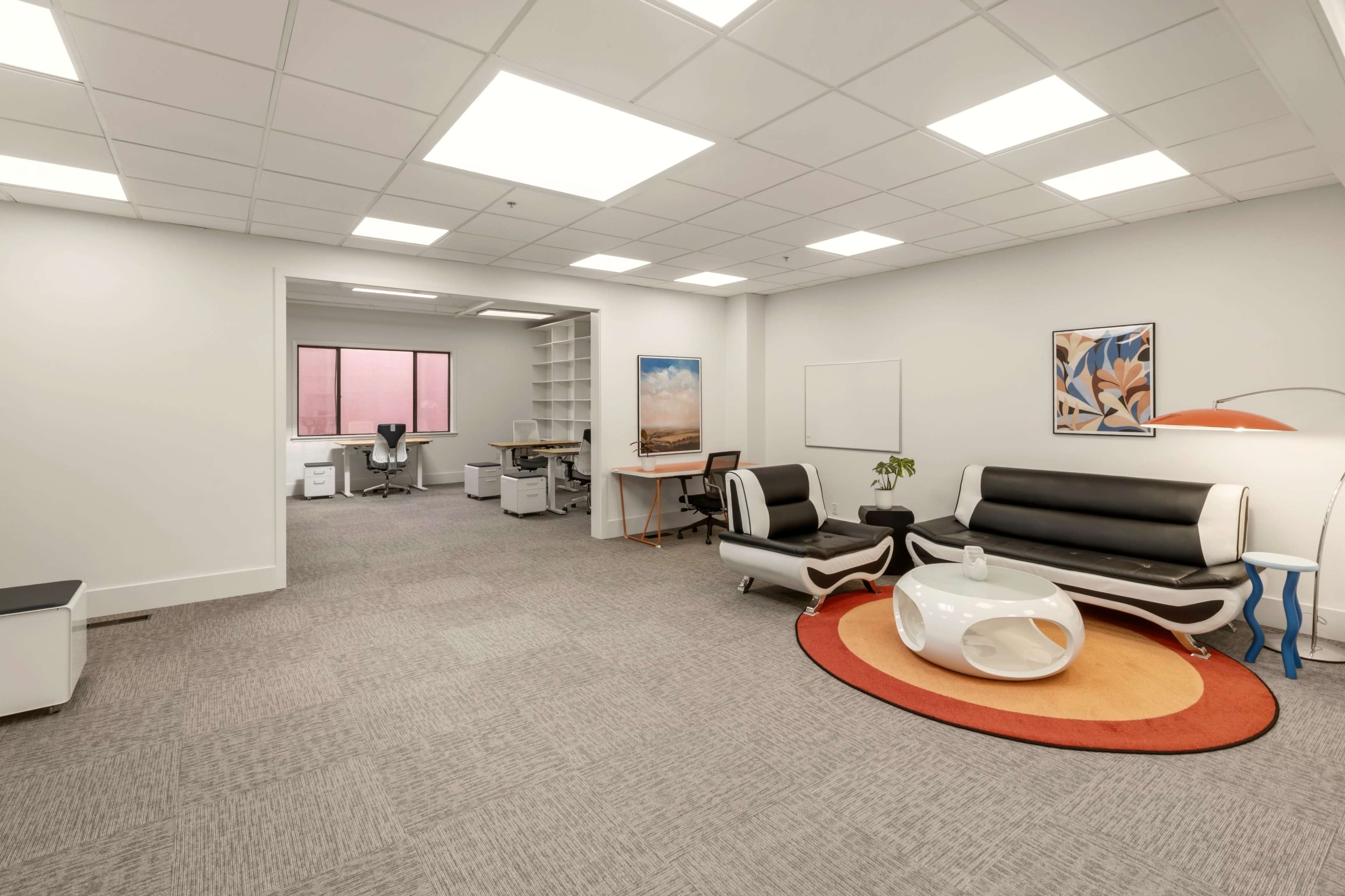 The image shows a modern office space featuring a seating area with a black and white couch, a round coffee table, and a bright orange rug, alongside workstations in the background.