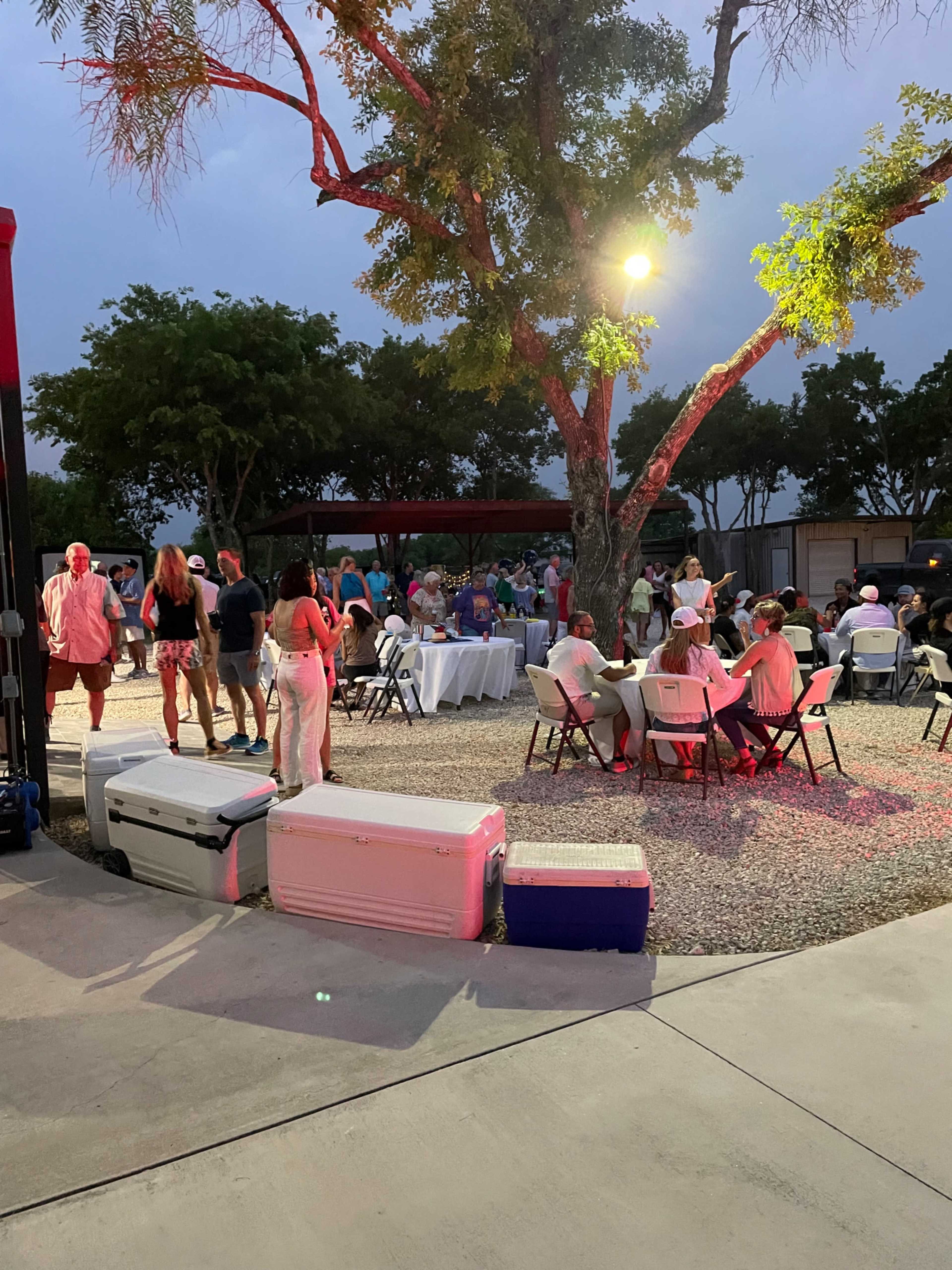 A gathering of people is taking place outdoors around tables, with coolers lined up on the ground, under soft lighting in a tree-lined area.