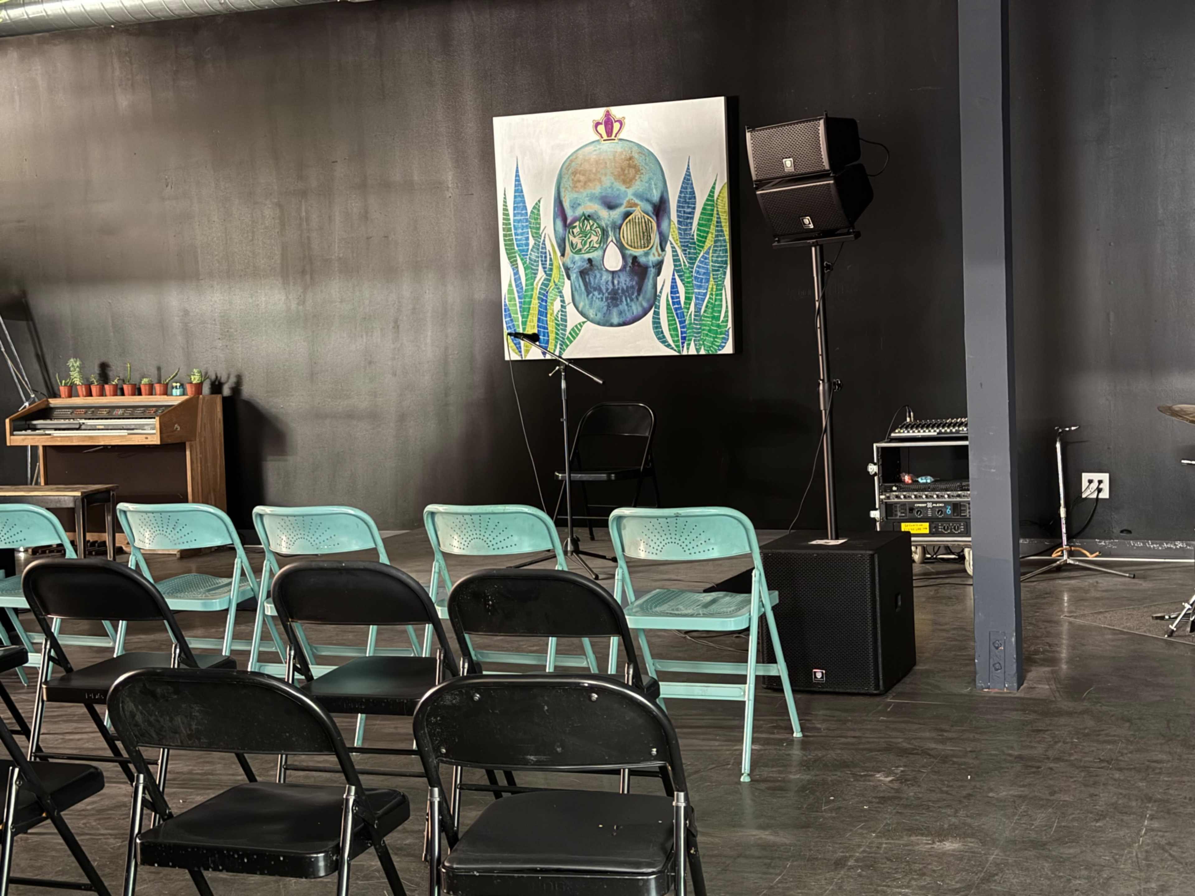 The image shows an indoor performance space with a large decorative skull painting on the wall, a microphone stand, and rows of black and turquoise chairs arranged for an audience.