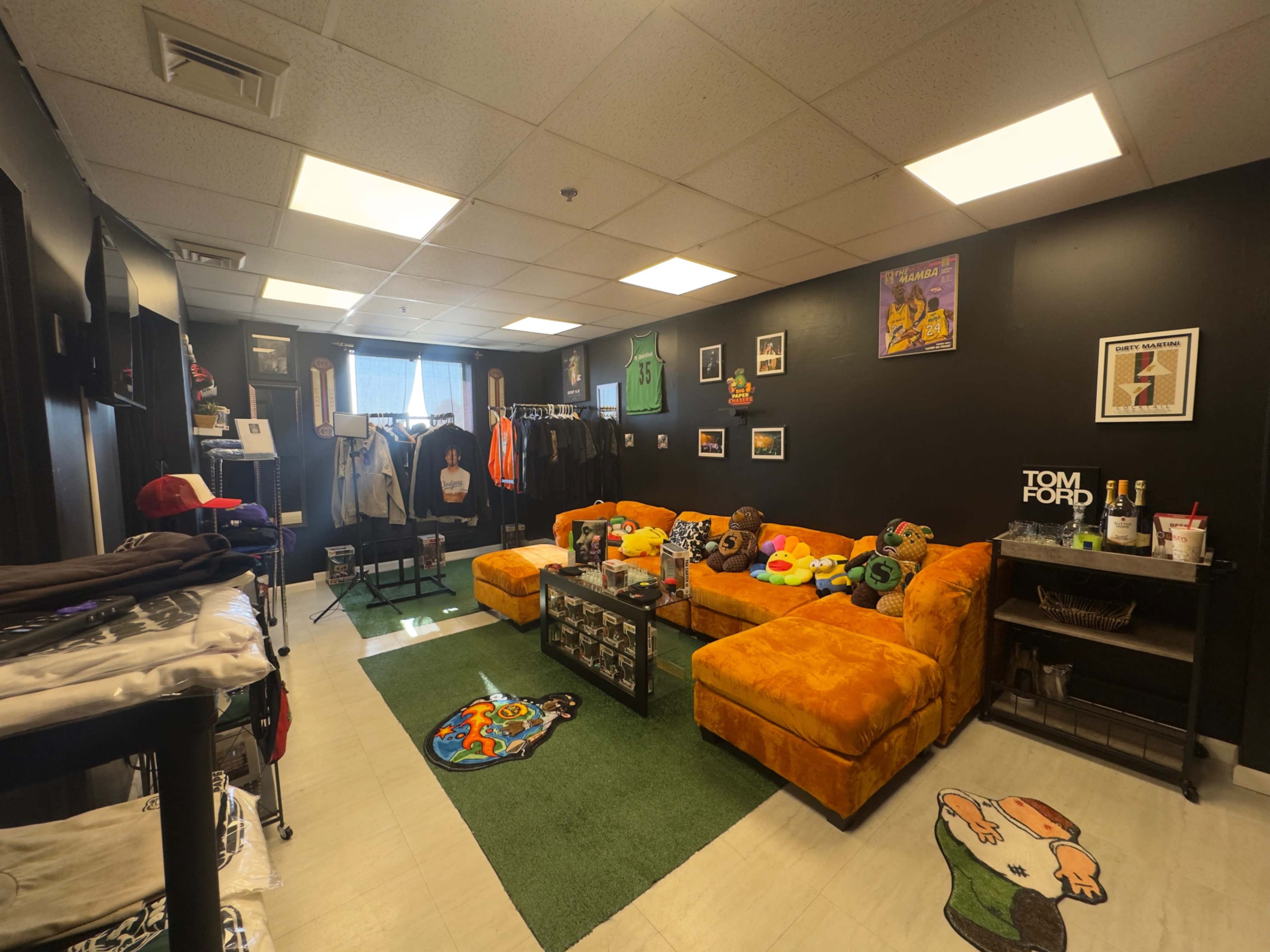 The room features a cozy seating area with orange couches adorned with various clothing items and decorative elements, set against a black wall displaying framed art and merchandise.