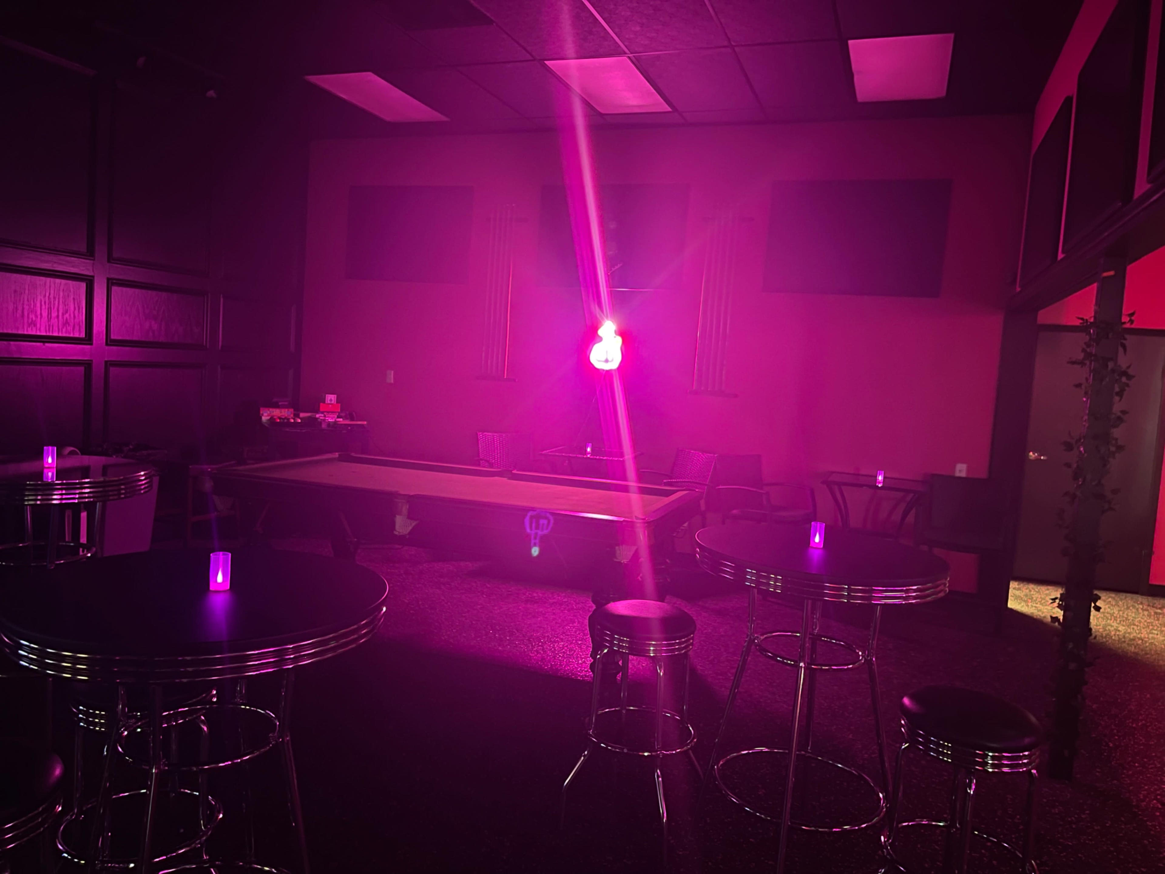 A dimly lit lounge features a pool table under a pink light, surrounded by small tables and chairs.