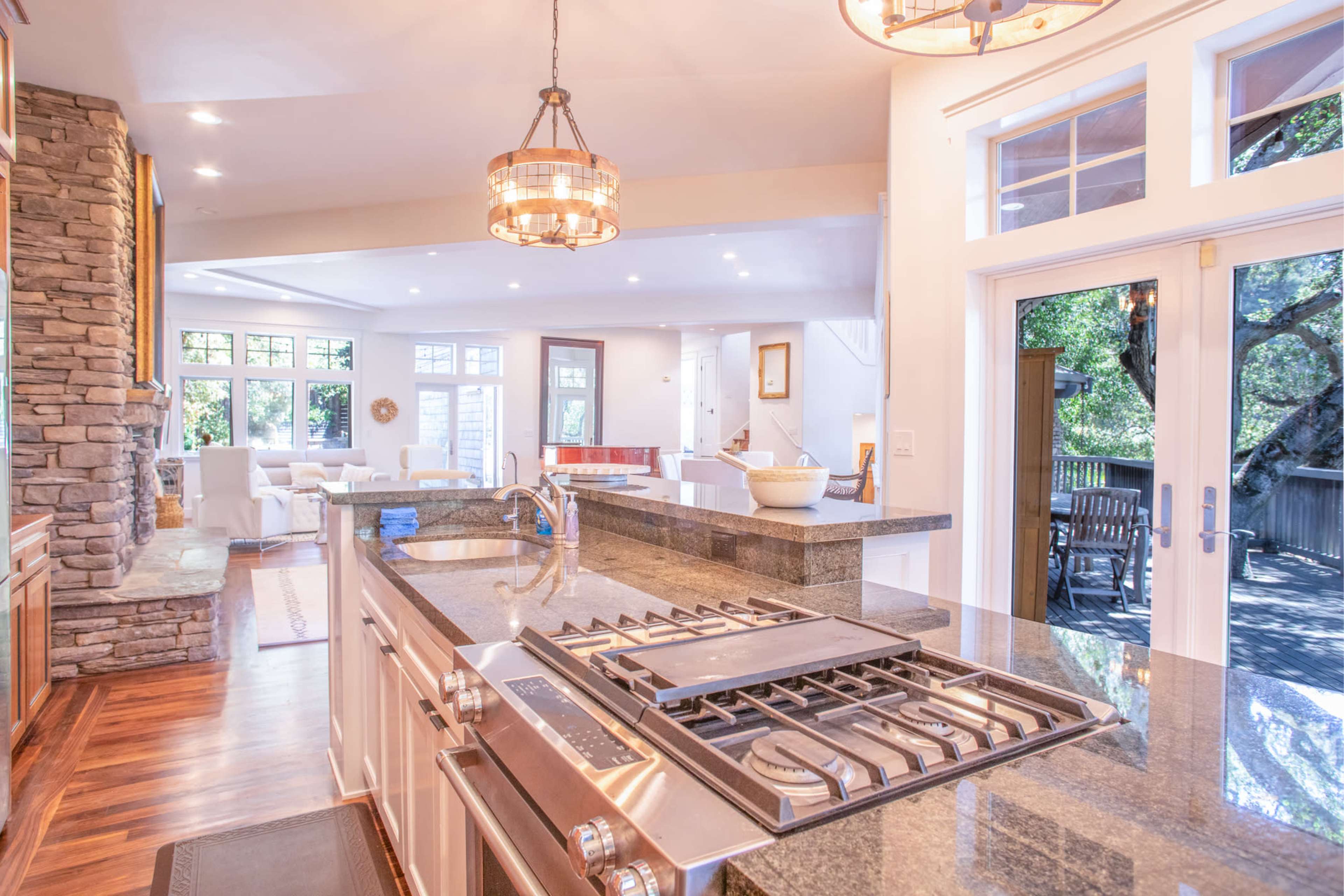 The image shows a modern kitchen with a granite countertop and a gas stove overlooking a spacious living area with large windows.