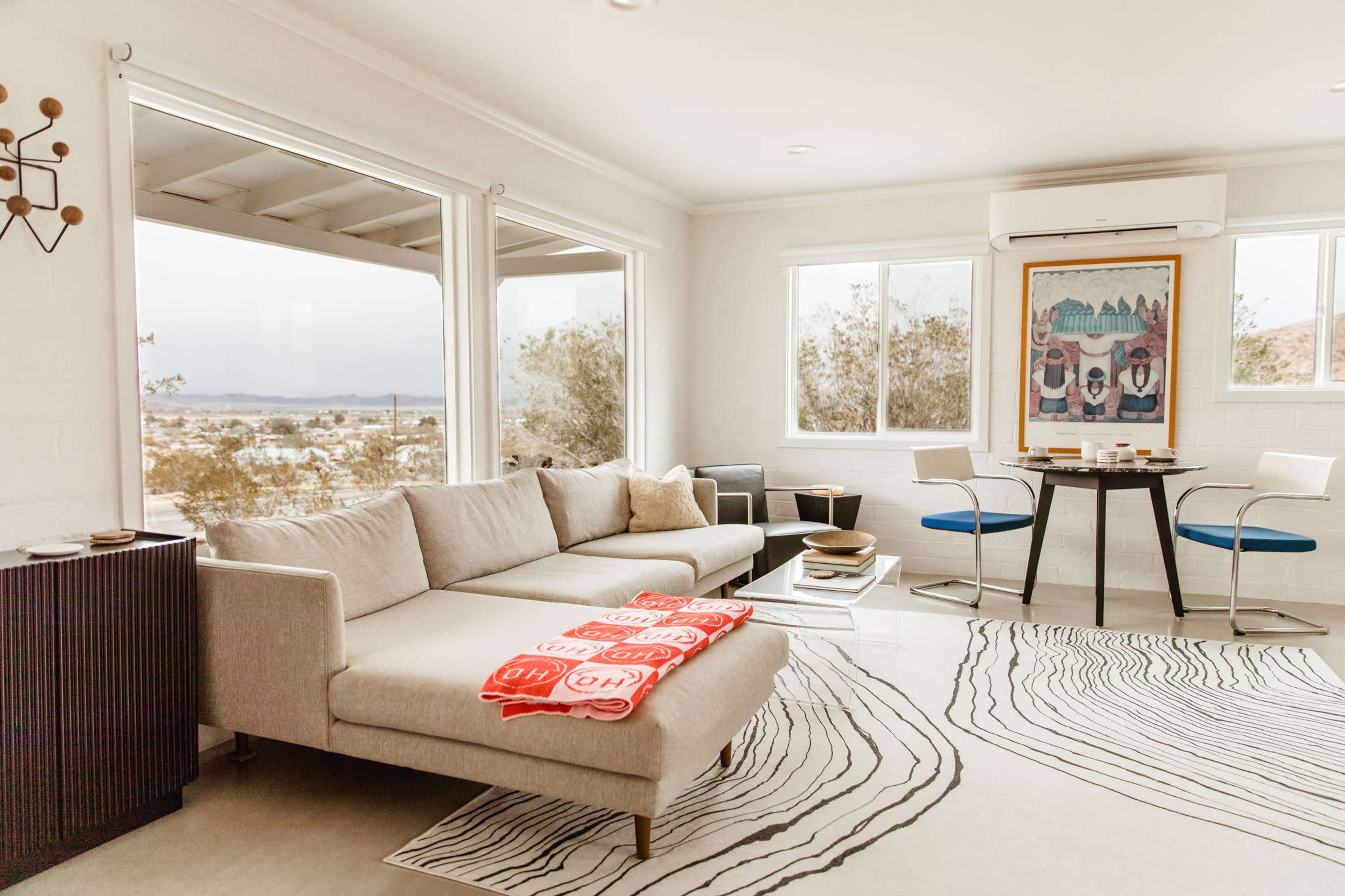 A modern living room features a sectional sofa, a circular dining table with chairs, and large windows offering a view of the desert landscape.