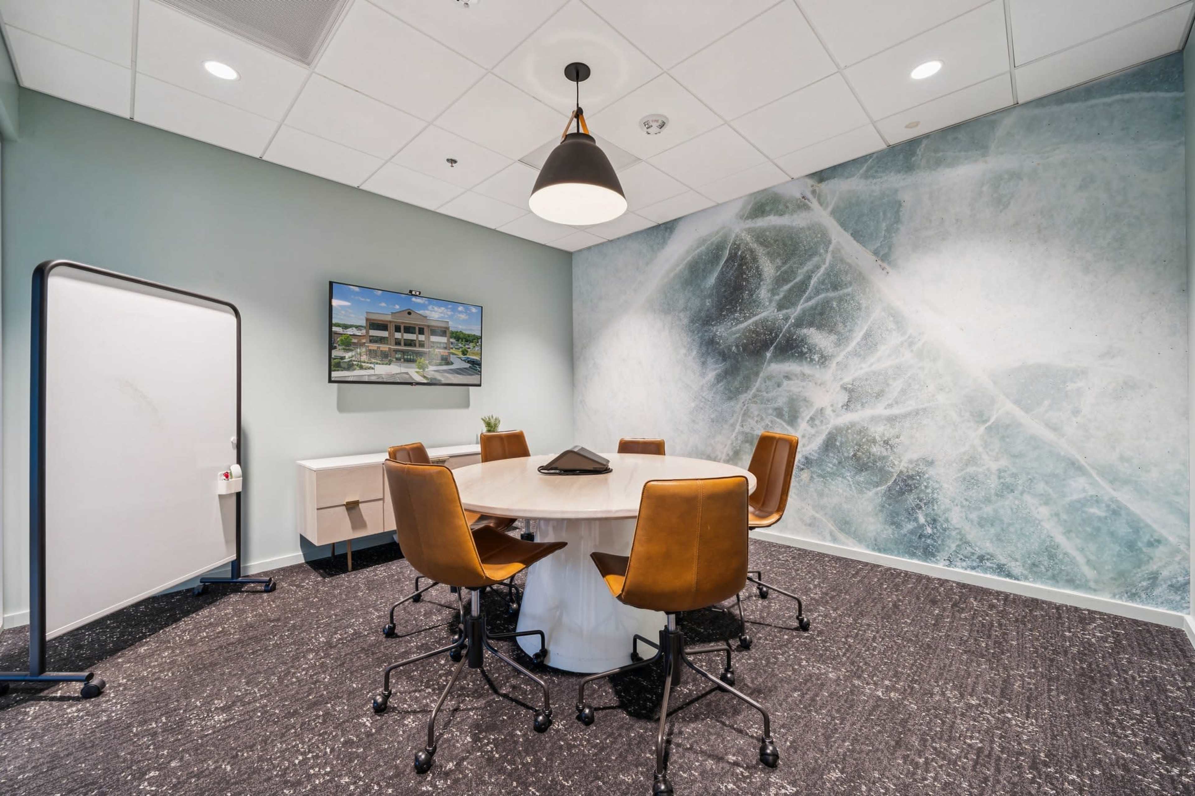 A modern conference room features a round table surrounded by brown leather chairs, with a large screen displaying an image on the wall, and a textured mural in the background.