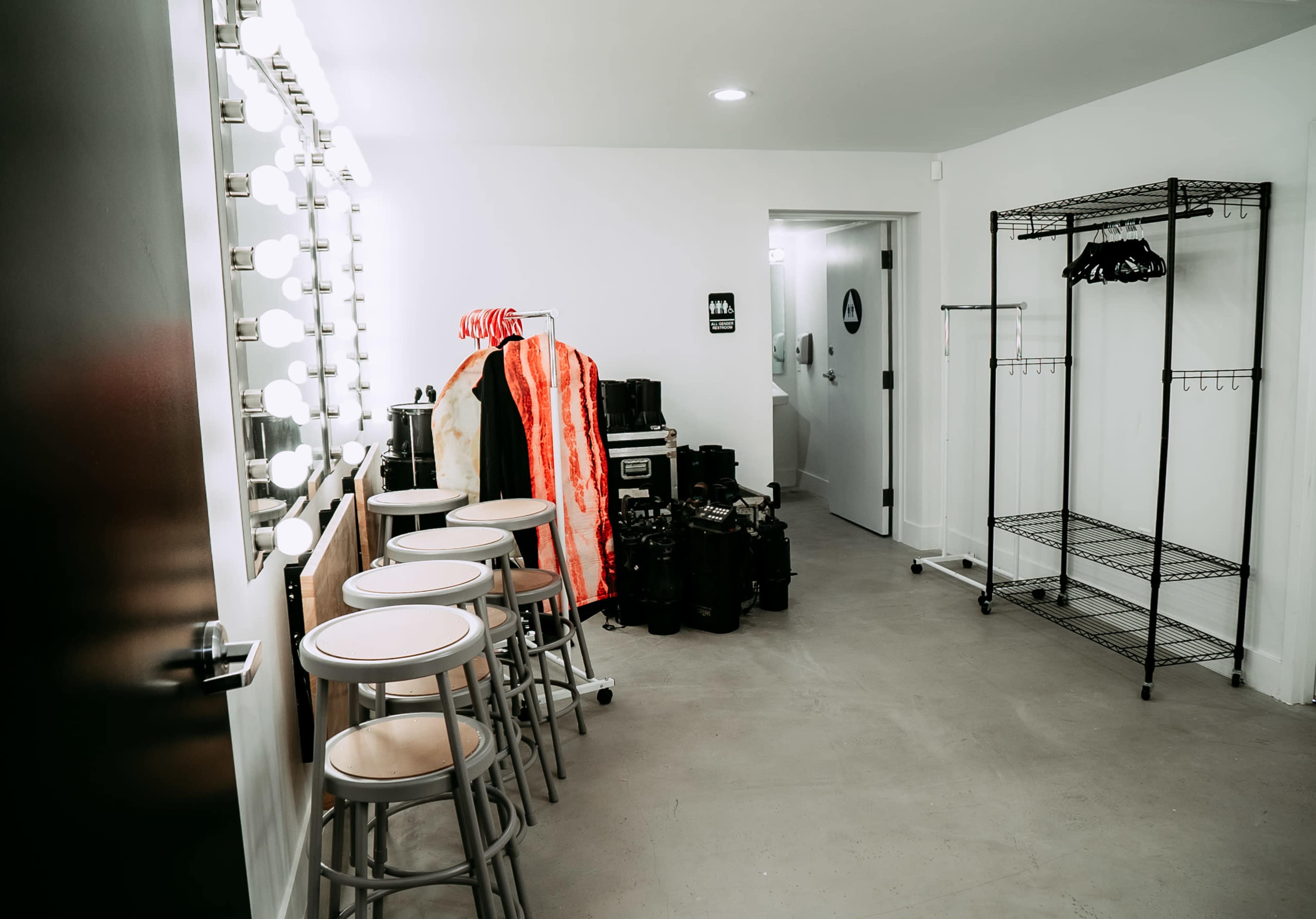 The image shows a well-lit dressing room with three stools, a clothing rack, and various equipment arranged against the white walls.
