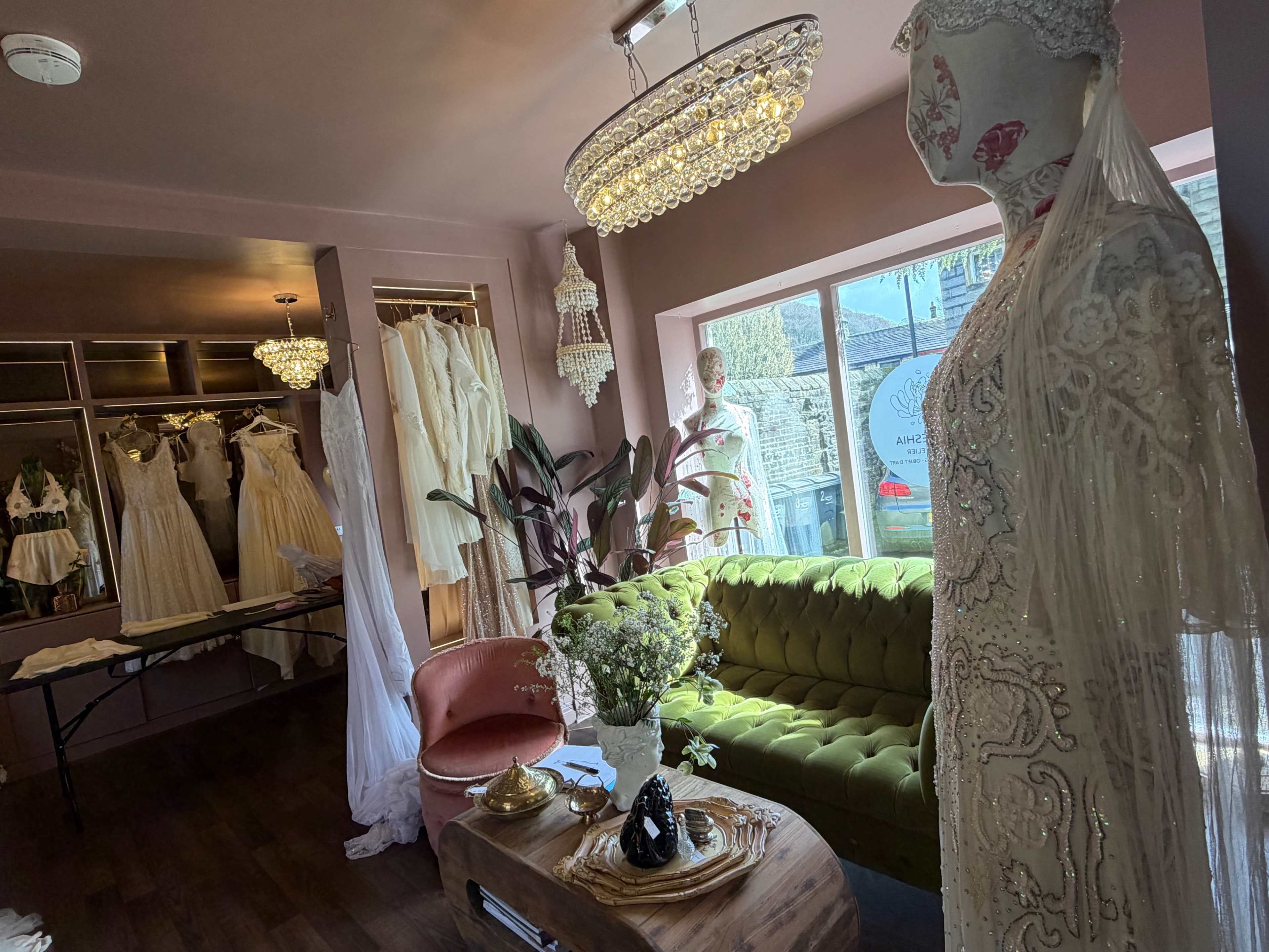 A bridal boutique features various wedding dresses displayed on hangers, with a green velvet sofa and a chandelier illuminating the room.