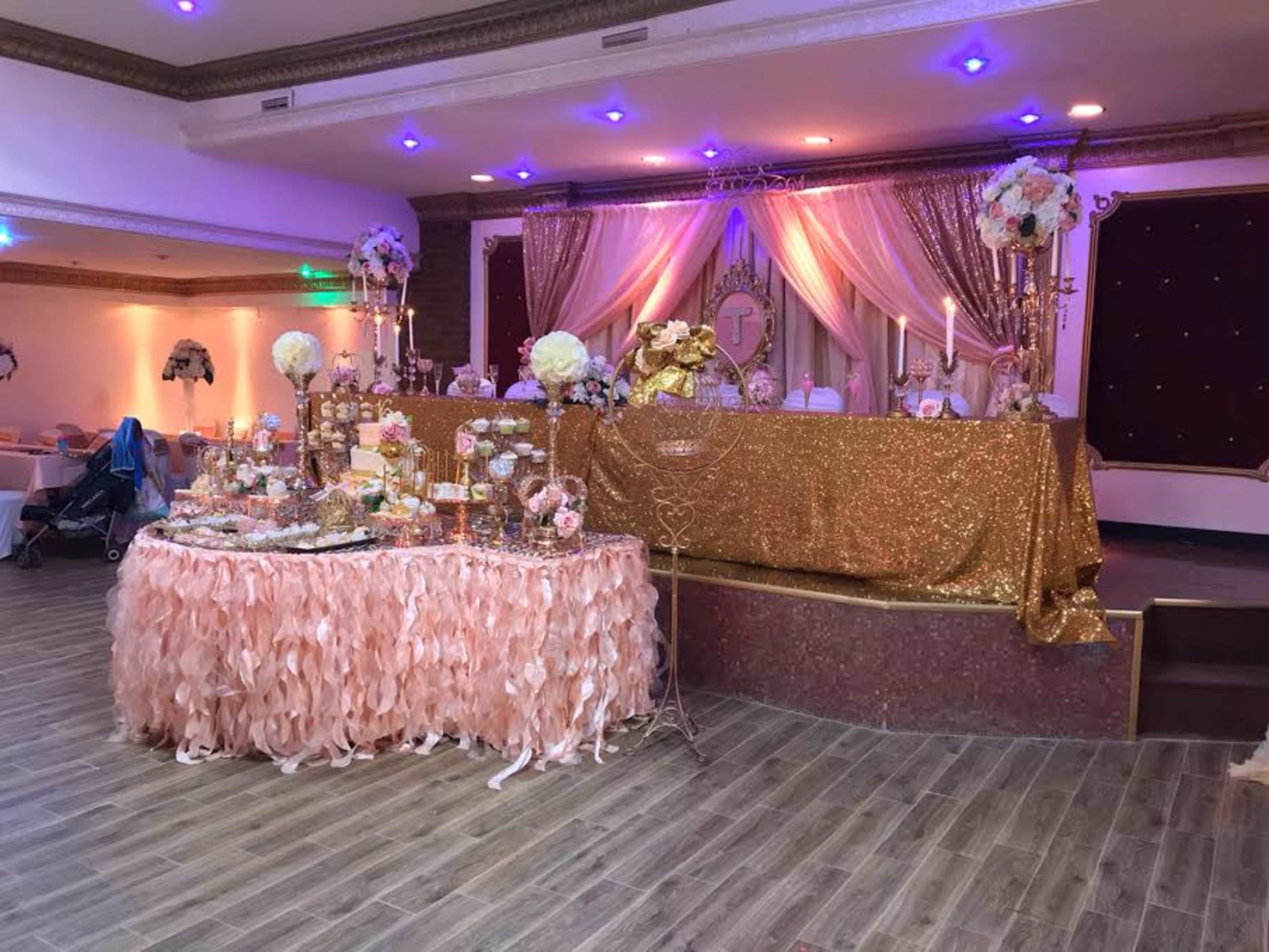 A decorated event space features a sparkling golden tablecloth backdrop with floral arrangements and a dessert table adorned with various sweet treats.