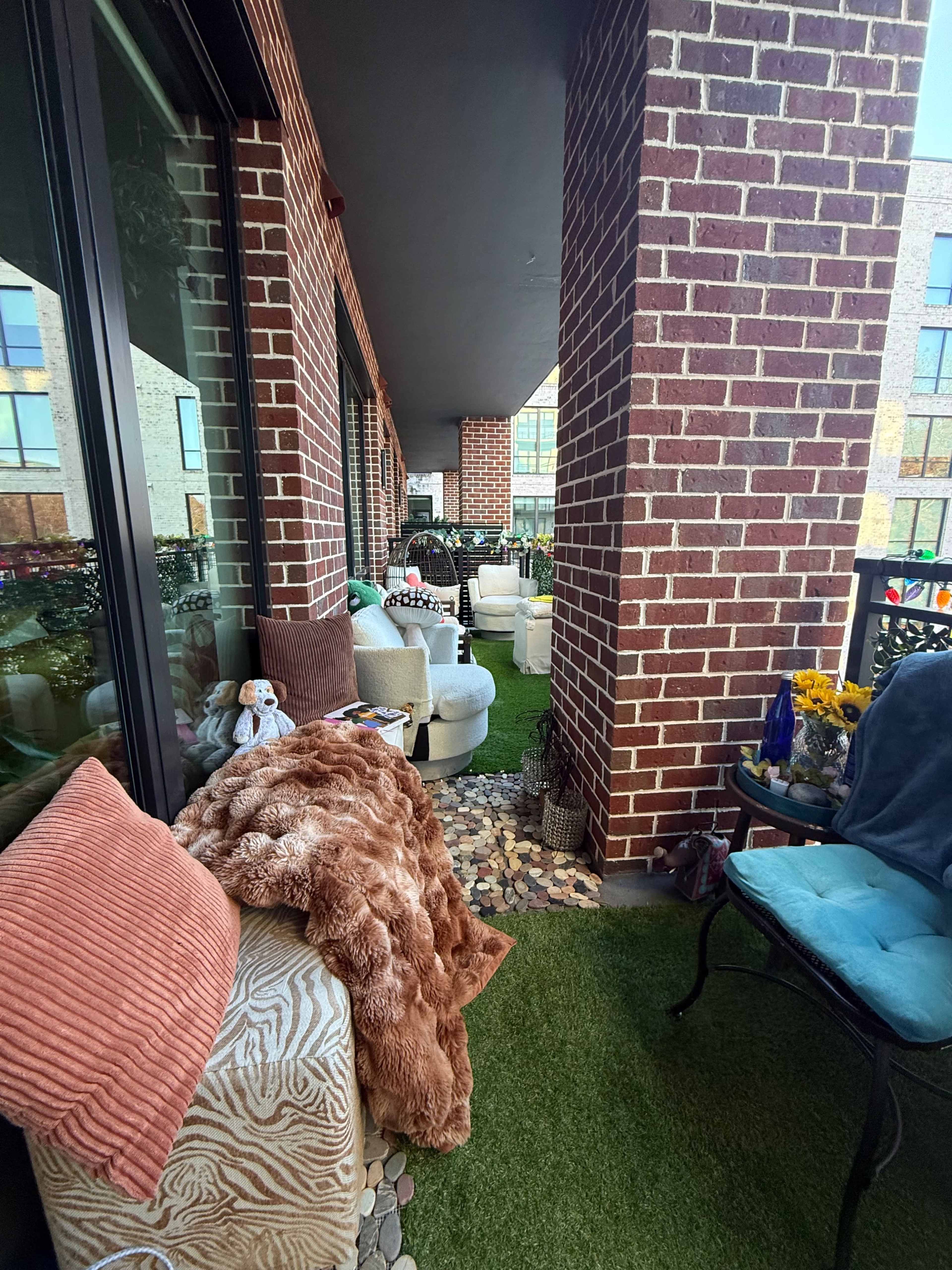 The image depicts a cozy outdoor balcony space furnished with various seating options, potted plants, and decorative pillows, bordered by brick columns.