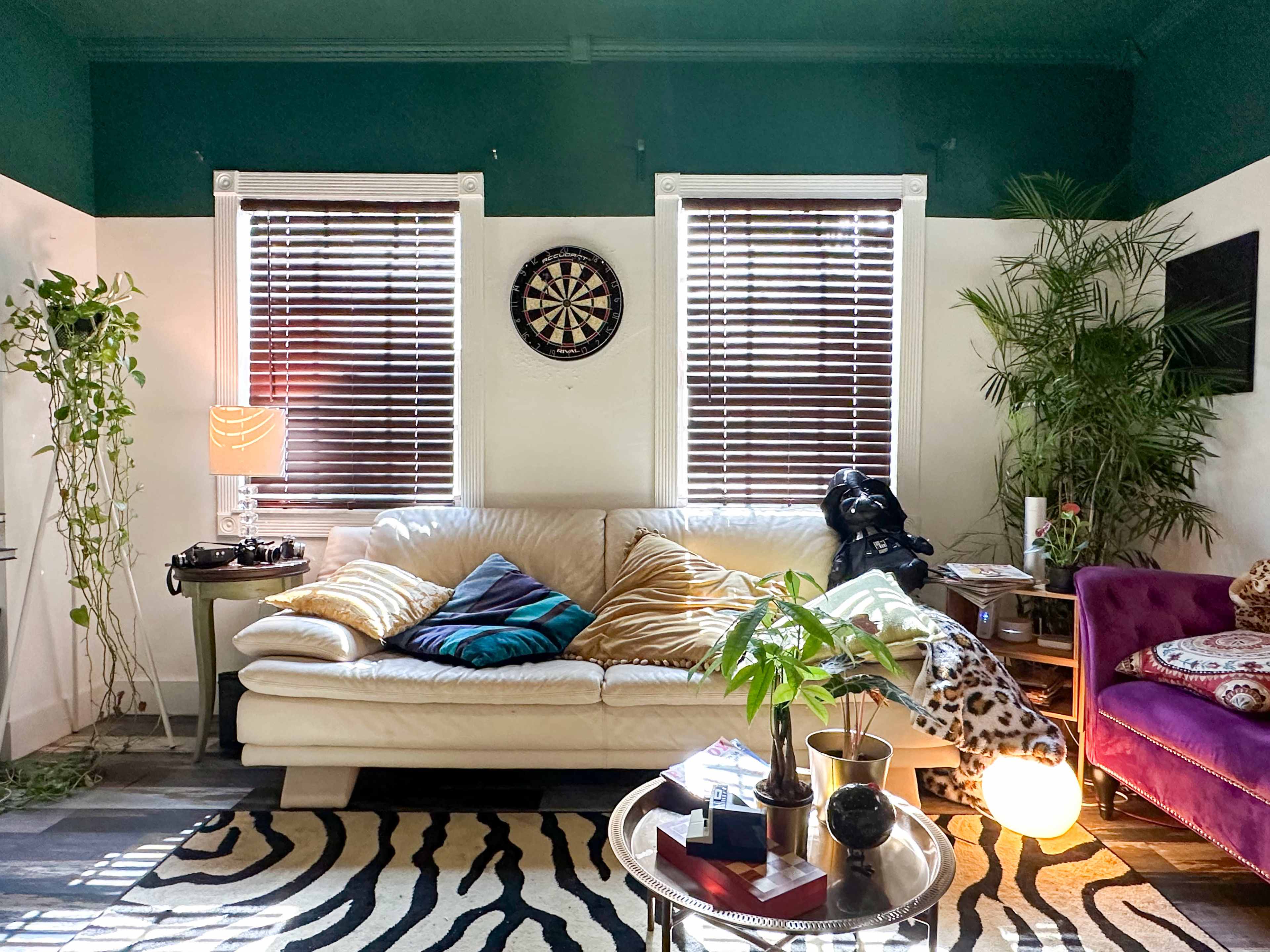 The scene features a living room with a white sofa adorned with colorful pillows, surrounded by plants and decorative furniture, including a dartboard on the wall and a record player nearby.