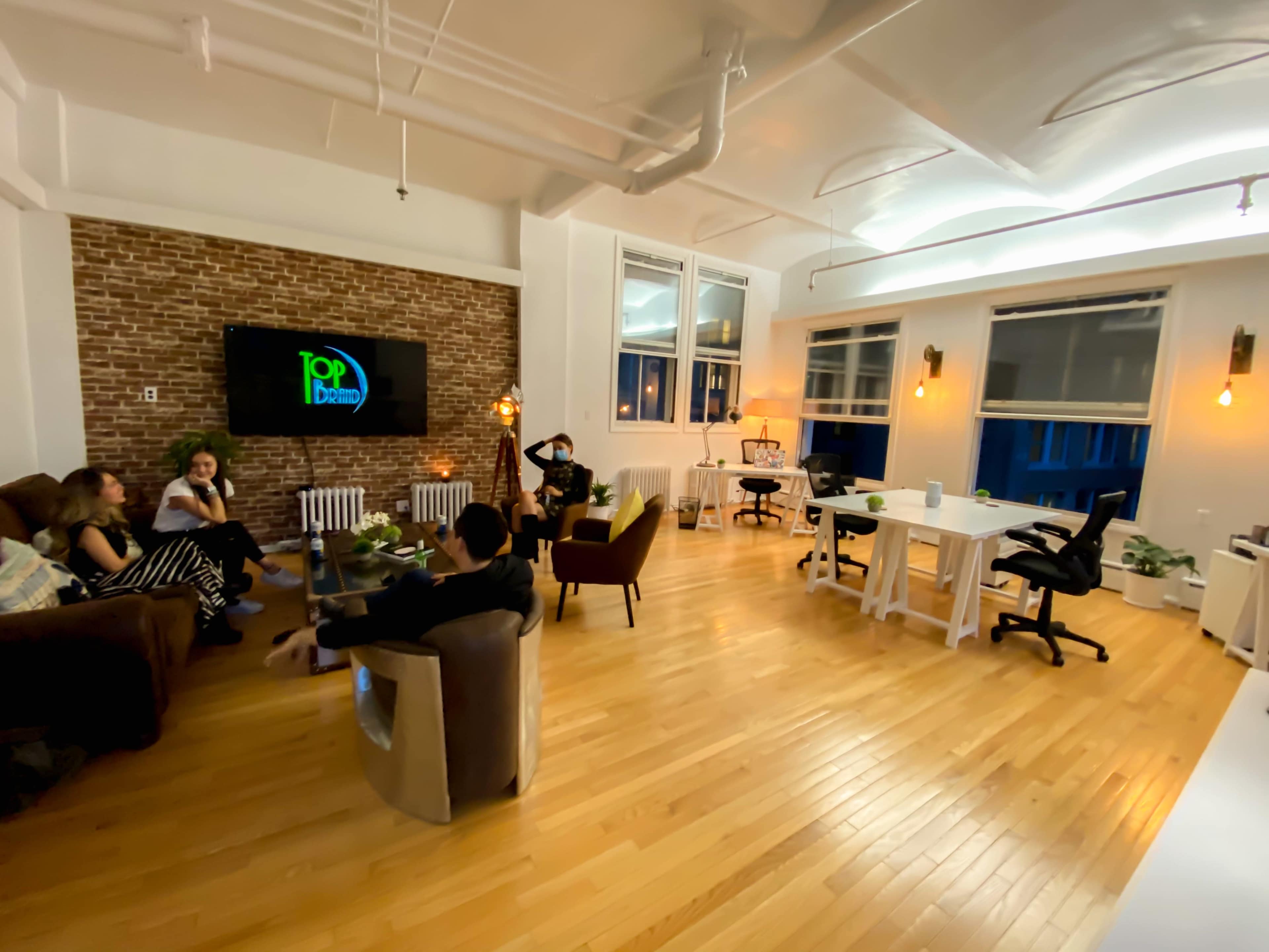 A modern co-working space features a large wooden floor, a brick accent wall, and several seating areas with people engaged in conversation.