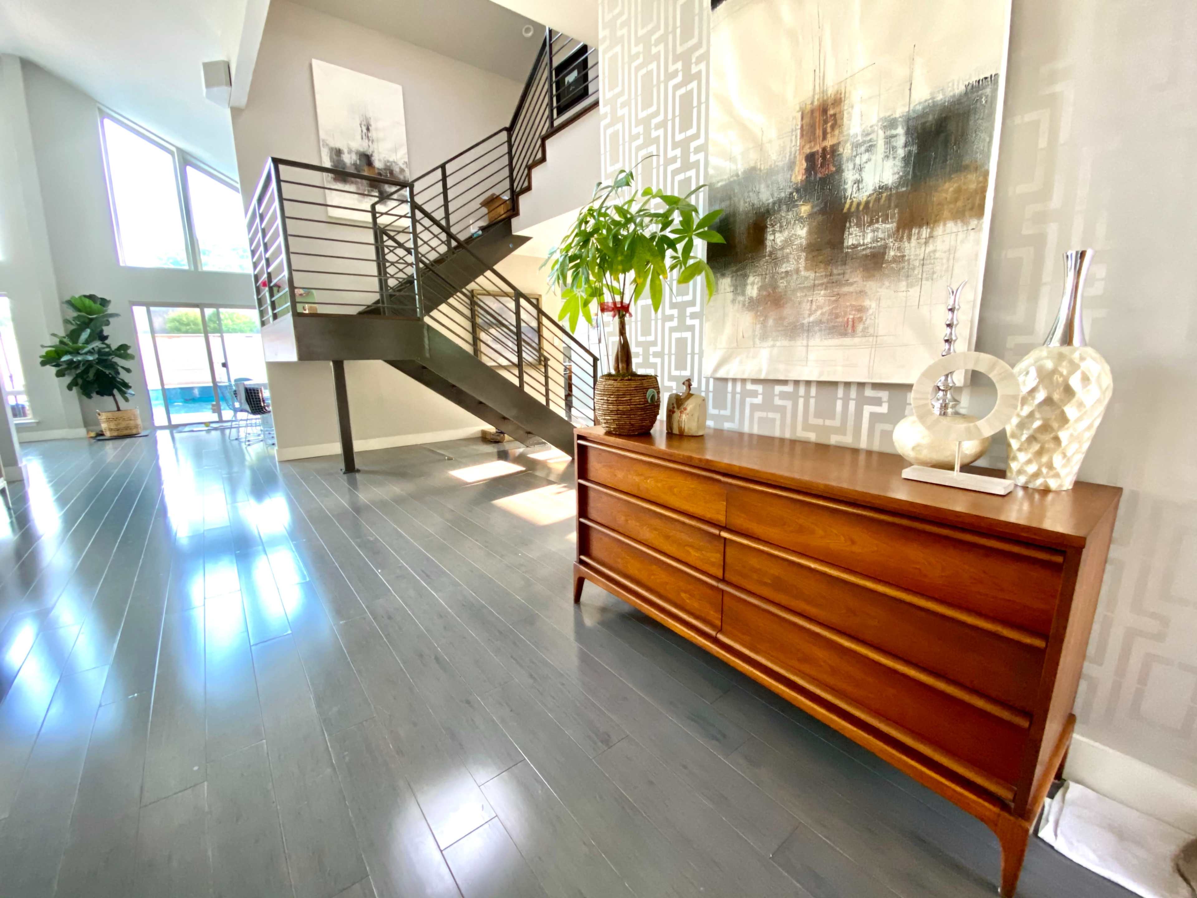 A spacious entryway features a wooden dresser, a potted plant, and a staircase leading to an upper level, with large windows allowing natural light to fill the space.