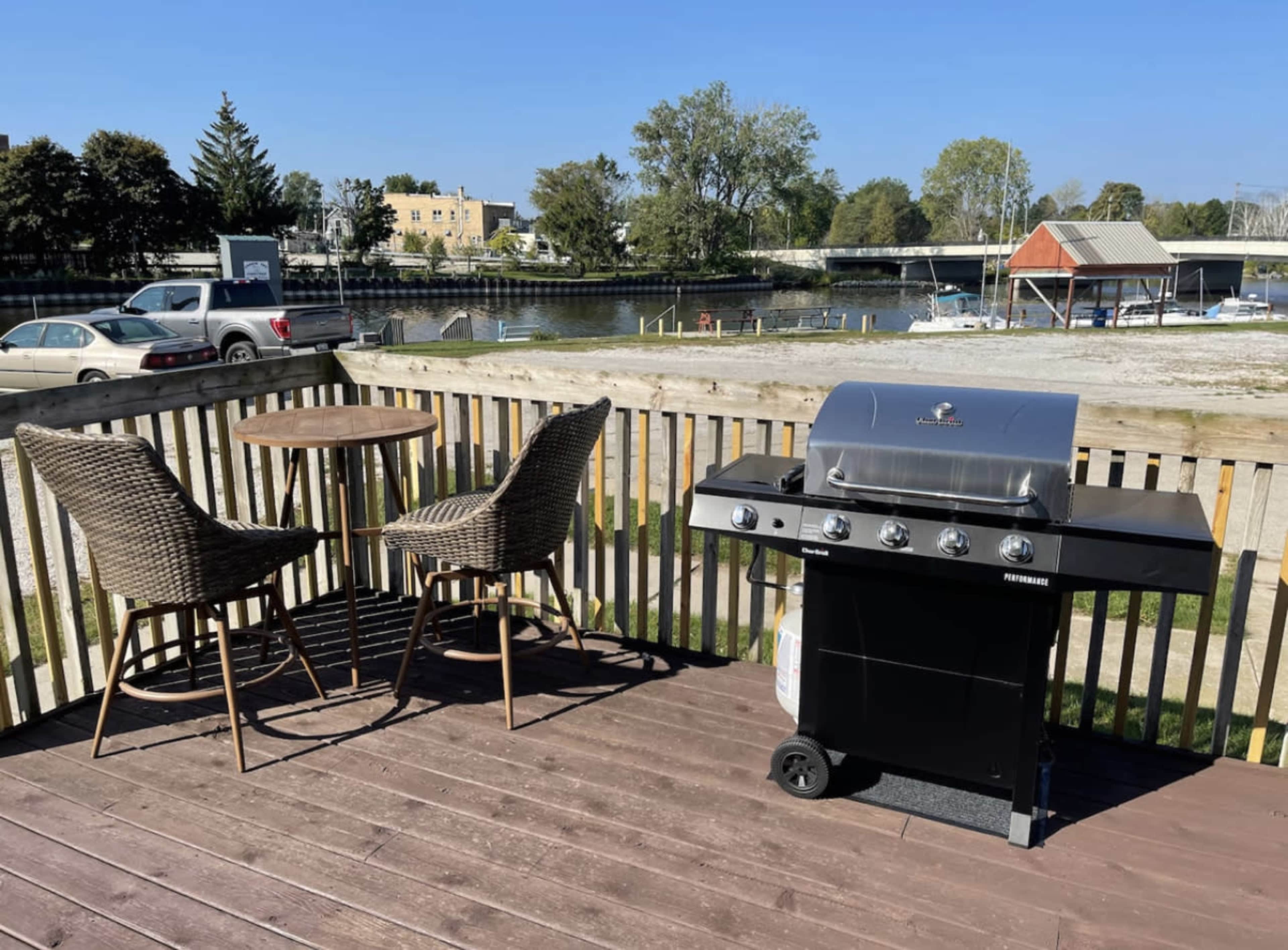 A gas grill is positioned on a wooden deck next to a round table and two wicker chairs, overlooking a waterway with boats and trees in the background.