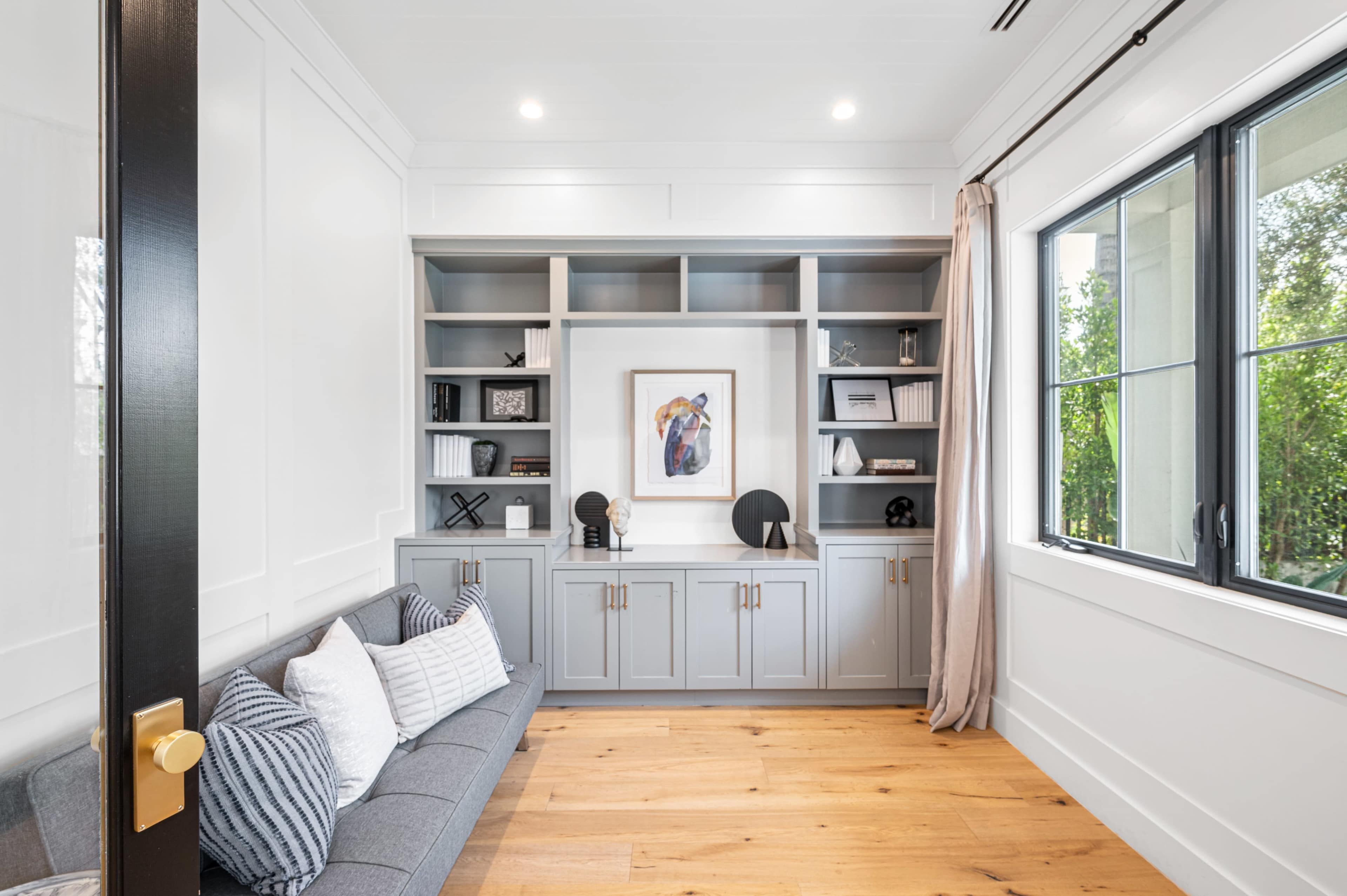 A neatly organized room features a built-in shelving unit filled with decor, with a gray couch and large window allowing natural light.