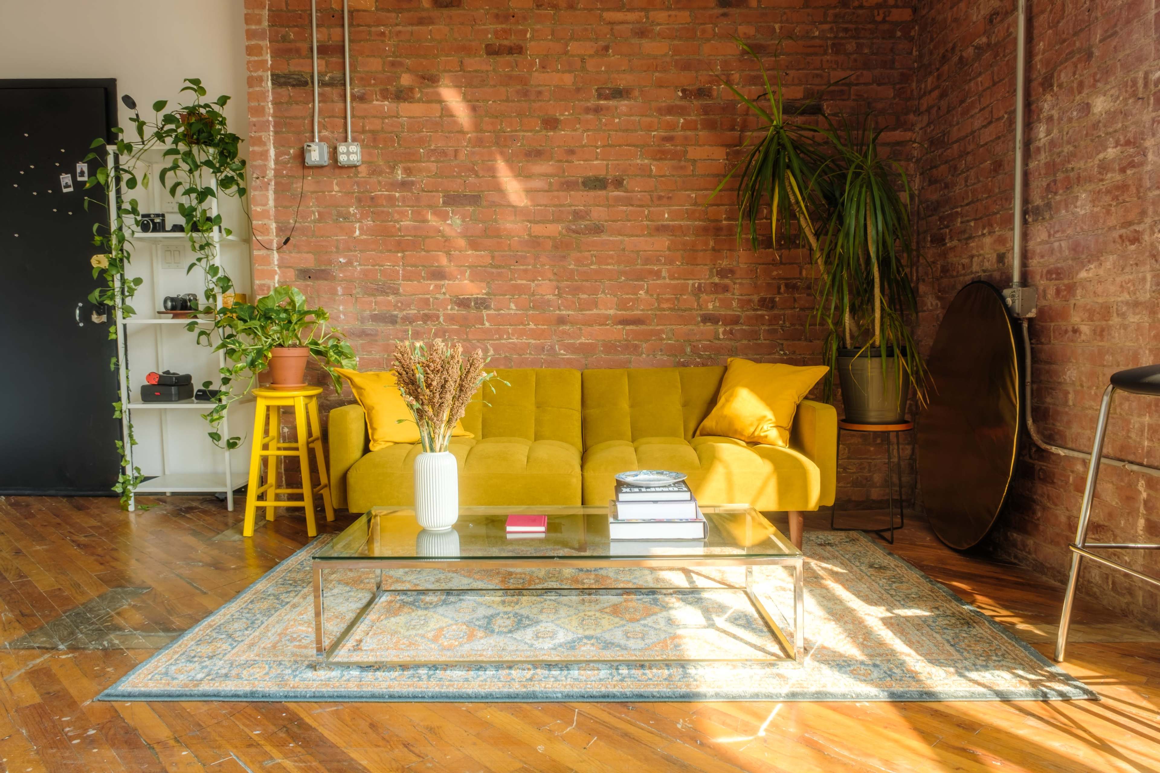 The image shows a cozy living area with a yellow sofa, a glass coffee table, a patterned rug, and exposed brick walls.