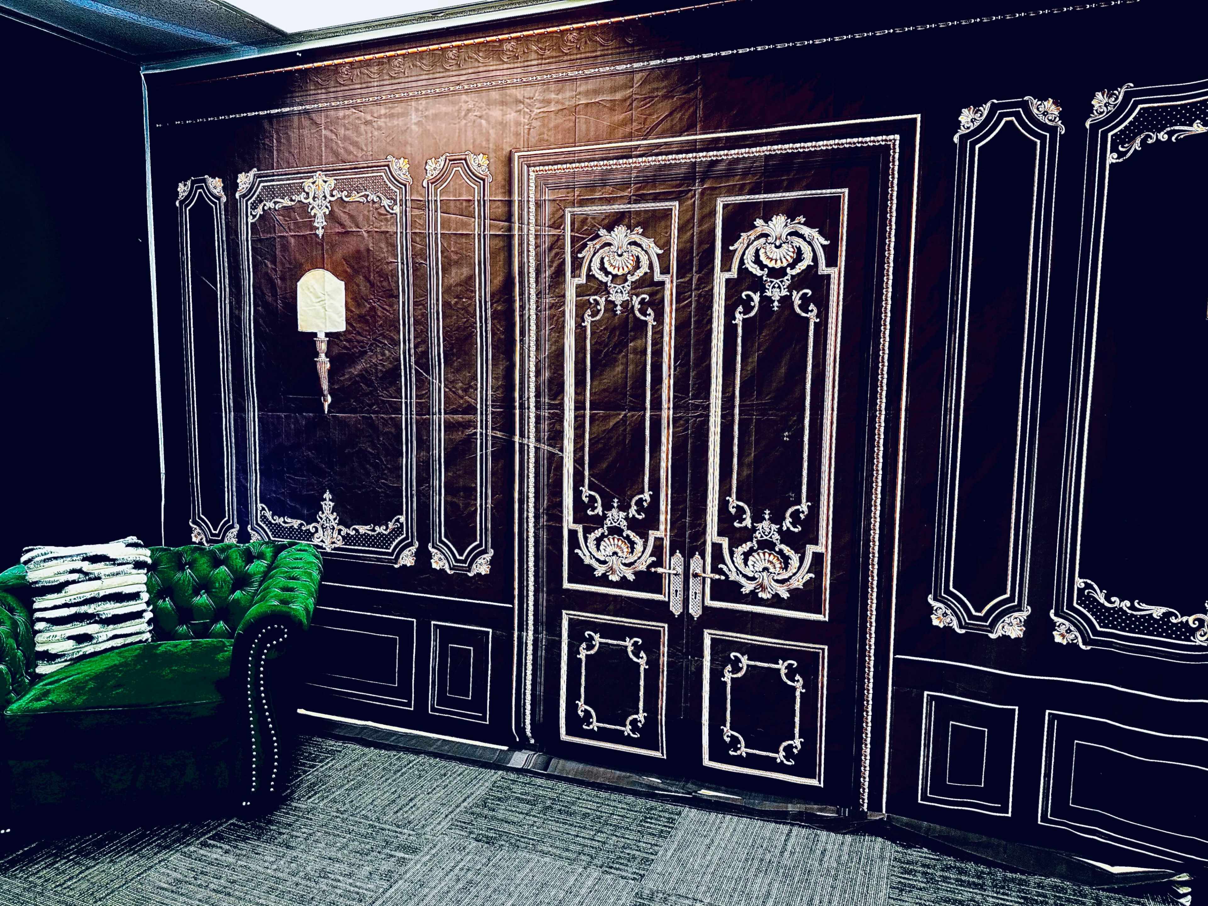 A room with a black and white ornate wallpaper featuring a door design, alongside a green velvet couch.