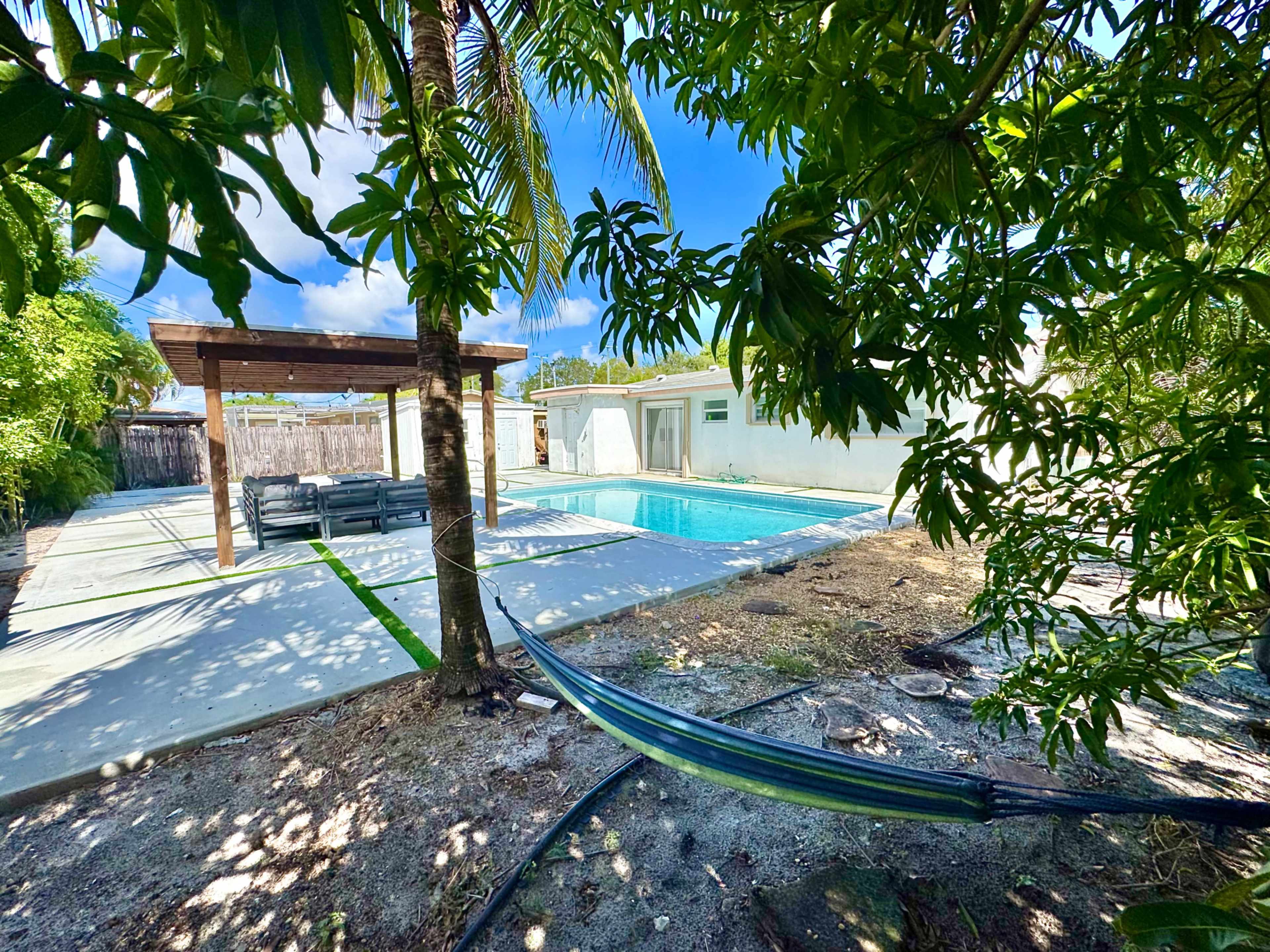 The image shows a backyard with a swimming pool, a covered seating area, and a hammock between palm trees.